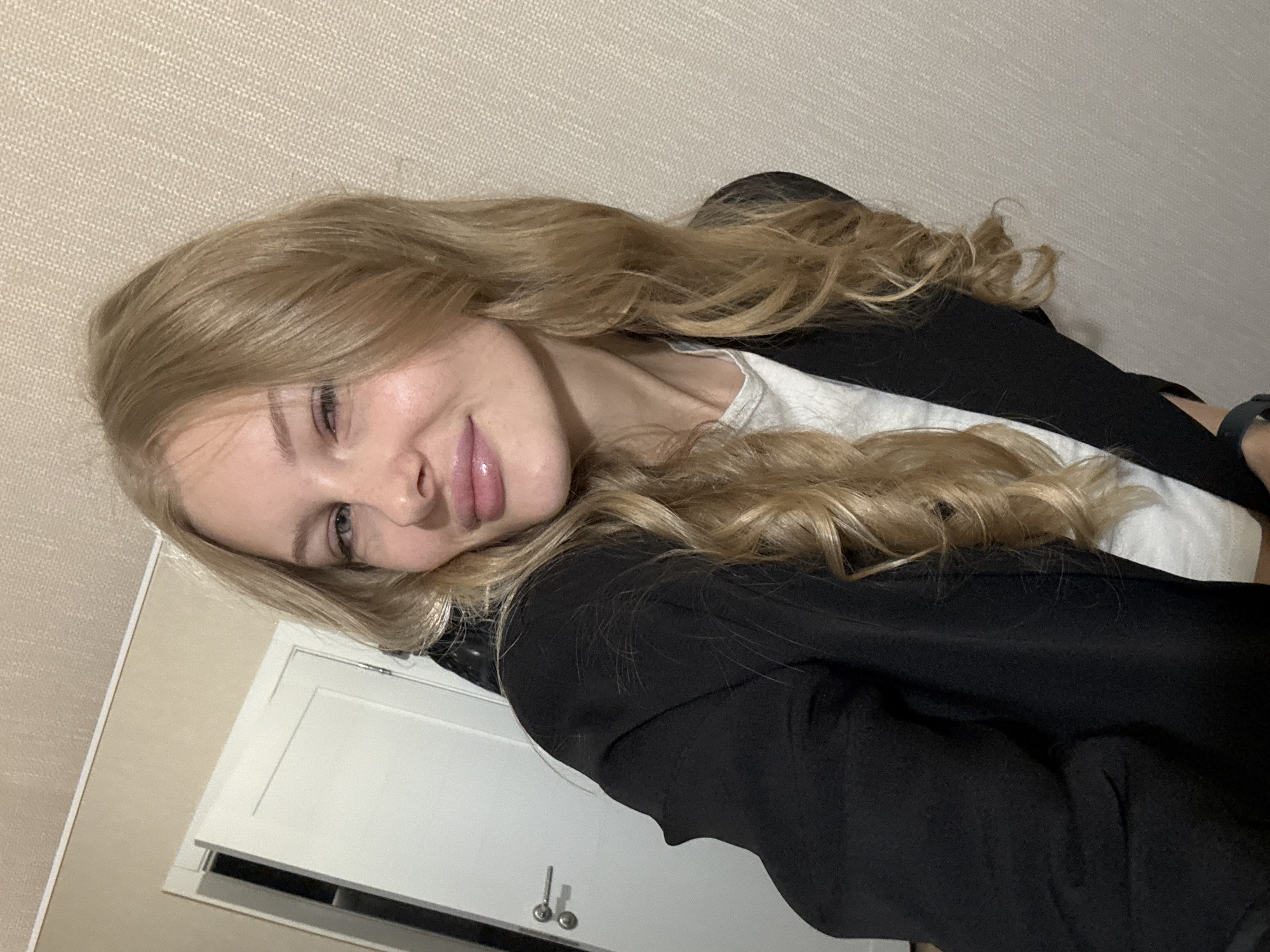 The image shows a young woman with blonde hair, wearing a black blazer over a white t-shirt. She is indoors, with a textured wall behind her and a white door with silver handles visible on the left. The scene appears to be an interior residential or office space. There is no visible indication of the city of Perm, Russia.
