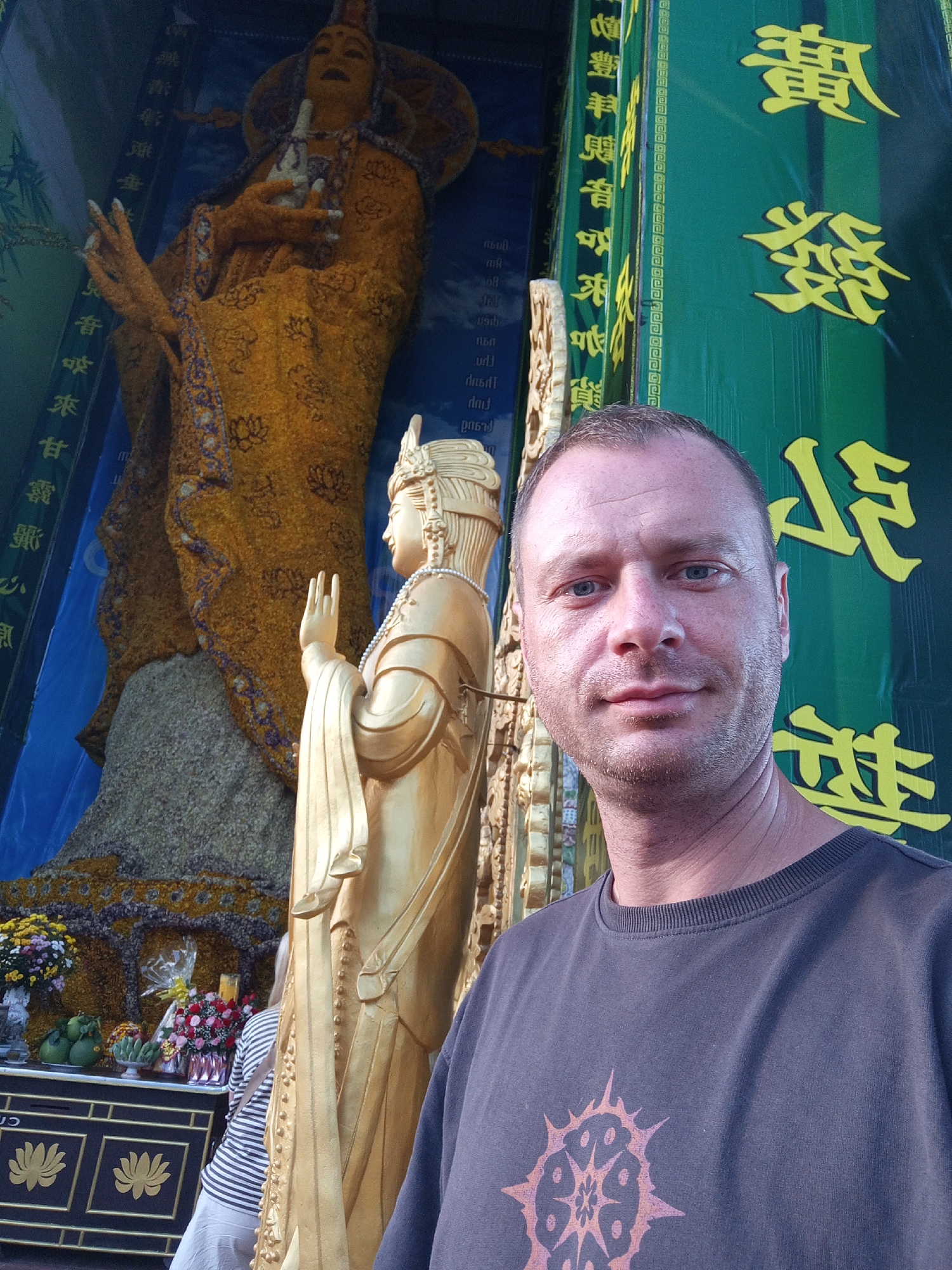 In Xuân Thành, Vietnam, a man is photographed in front of a large, ornate golden statue of a deity. The statue appears to be adorned with floral arrangements. Behind the man and the statue, there are large green banners with yellow Chinese characters. To the left of the statue, there is another, larger golden statue partially visible, also with intricate detailing. The lower portion of the image shows a display of fruits and flowers on a table in front of a decorative cabinet. The man is looking directly at the camera, with a neutral expression. The lighting suggests an outdoor or well-lit indoor environment.