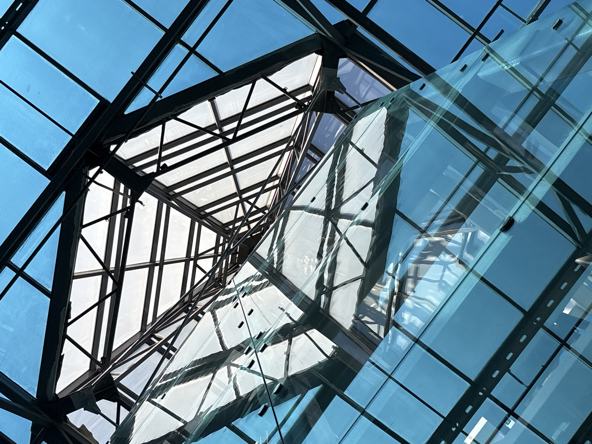 The image shows an interior view of a modern building's atrium, looking upwards. The structure is characterized by a complex geometric design of dark metal beams supporting large panes of translucent blue and clear glass. The glass panels form a vast, angular roof and enclose a central, curving glass structure that appears to be a walkway or elevator shaft. The clear glass panels reveal the intricate framework above and reflect the blue glass and sky. The scene is devoid of any people or animals, focusing entirely on the architectural elements and the interplay of light and reflection. The context provided indicates the location is Saint Petersburg, Russia.