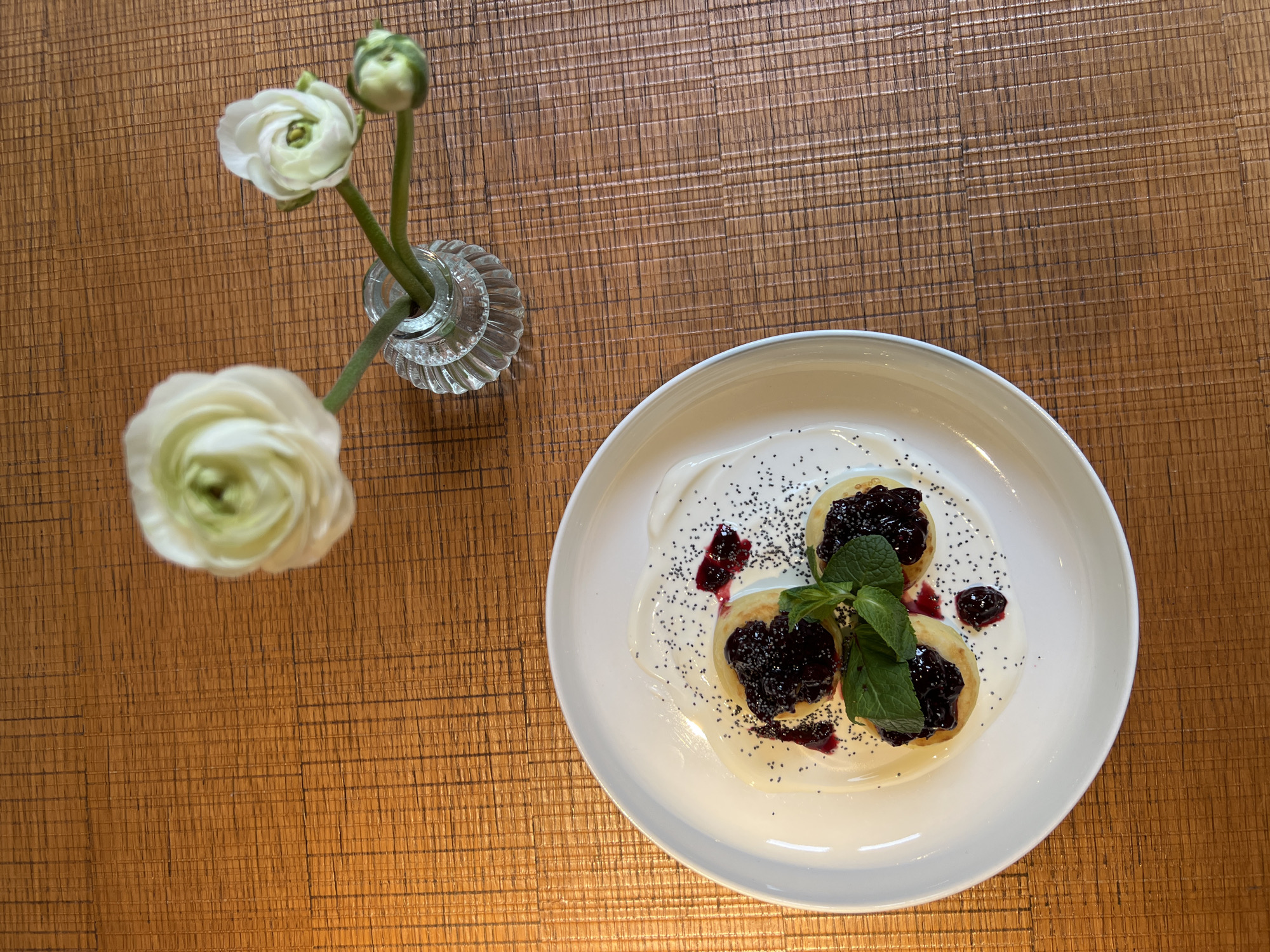 The image captures a scene on a wooden table, featuring a plate of what appears to be syrniki (cheese pancakes) topped with blueberry jam, a sprig of mint, and poppy seeds. The syrniki are arranged on a white plate with a light-colored sauce, possibly sour cream. Beside the plate is a small glass vase holding two white ranunculus flowers, one fully bloomed and another still in bud. The background reveals a textured, wood-grained tabletop, likely in a restaurant or cafe setting. While the image provides no explicit clues to the specific location in Saint Petersburg, Russia, the typical dish and flower arrangement suggest a refined, cozy ambiance, typical of eateries in the city.