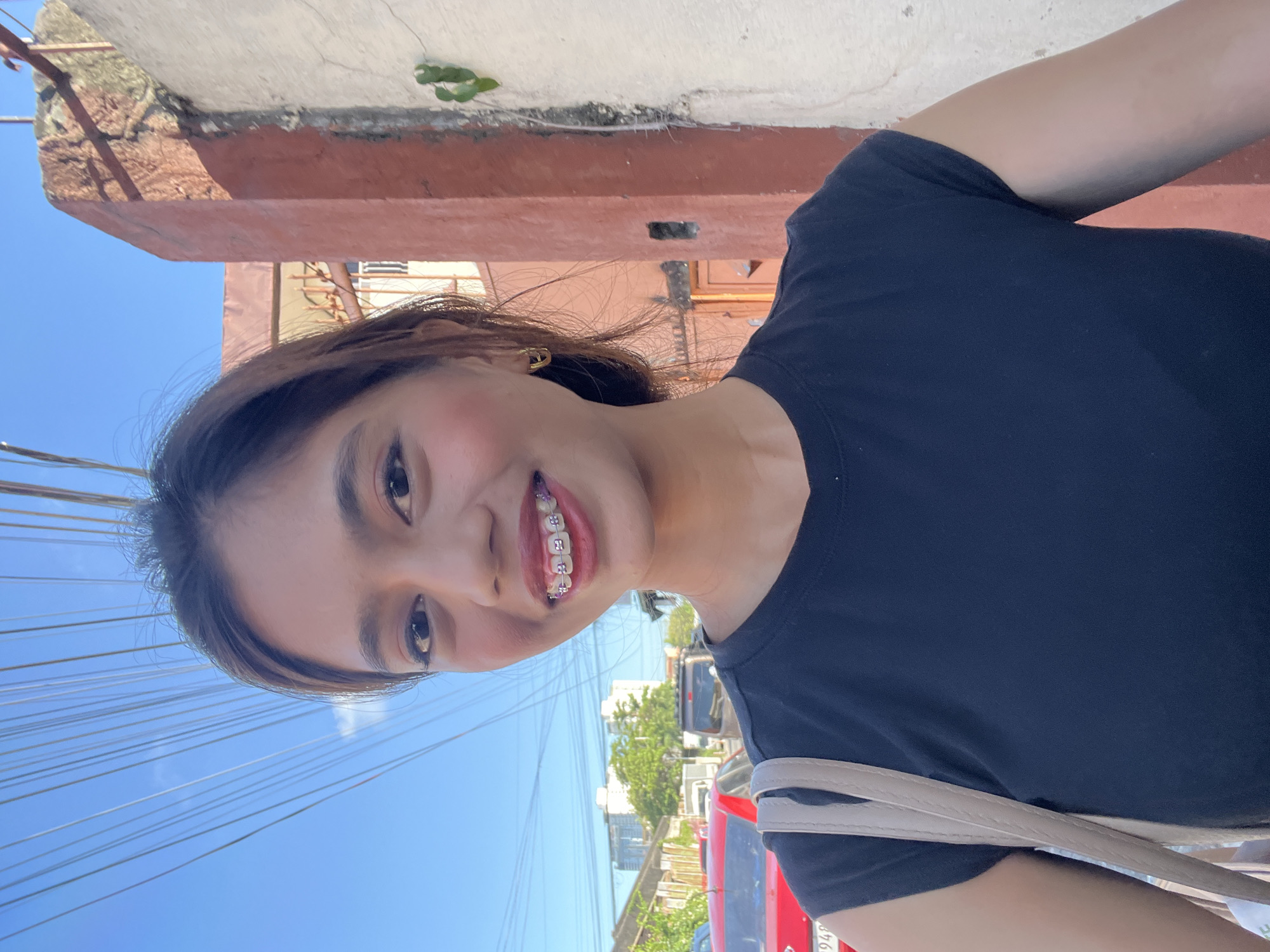 A young woman with braces smiles at the camera in this outdoor, daytime, selfie-style photograph. She is wearing a black t-shirt and a beige shoulder bag. Her dark hair is pulled back, and she has on light makeup, including pink blush and lip gloss. She is wearing a pair of gold hoop earrings. In the background, a bright blue sky is visible with numerous overhead wires. To the right of the woman is a rough, textured wall of a building, painted in shades of white and rust orange. Further in the background, there is a street scene with parked cars, including a red car and a dark van, and trees and buildings visible in the distance. The overall setting suggests an urban environment in Davao City, Philippines, likely a residential or commercial street.
