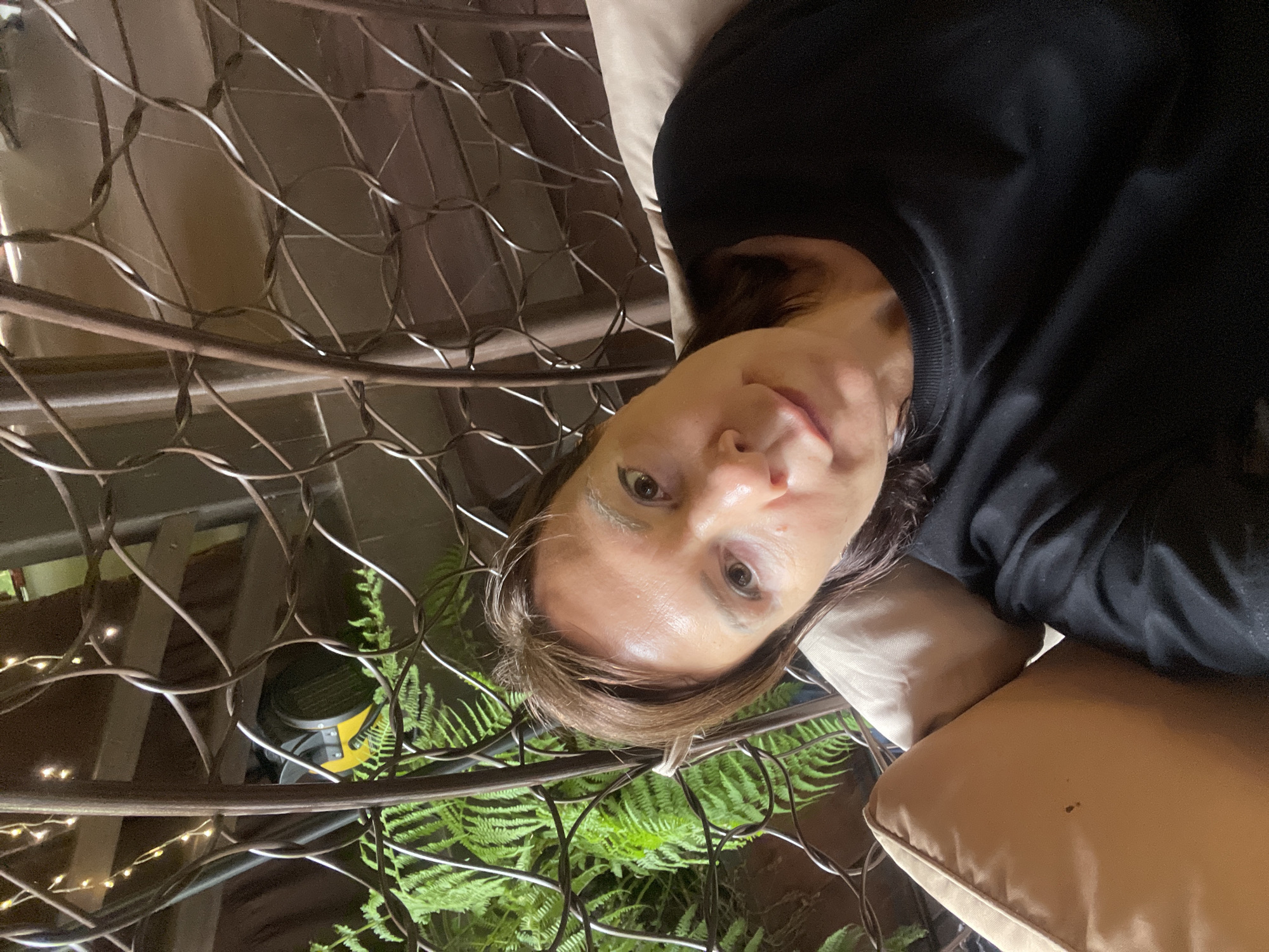 A selfie of a woman wearing a black long-sleeved shirt is positioned against a backdrop featuring a metal wire structure, possibly a cage or decorative element. Green ferns are visible behind the wire structure, along with indistinct lighting elements. A tan-colored cushion is partially visible, supporting the woman's head and shoulder. The woman appears to be in a resting or reclining position. The location is potentially Solokh-Aul, Russia, although there is no definitive visual confirmation within the image.