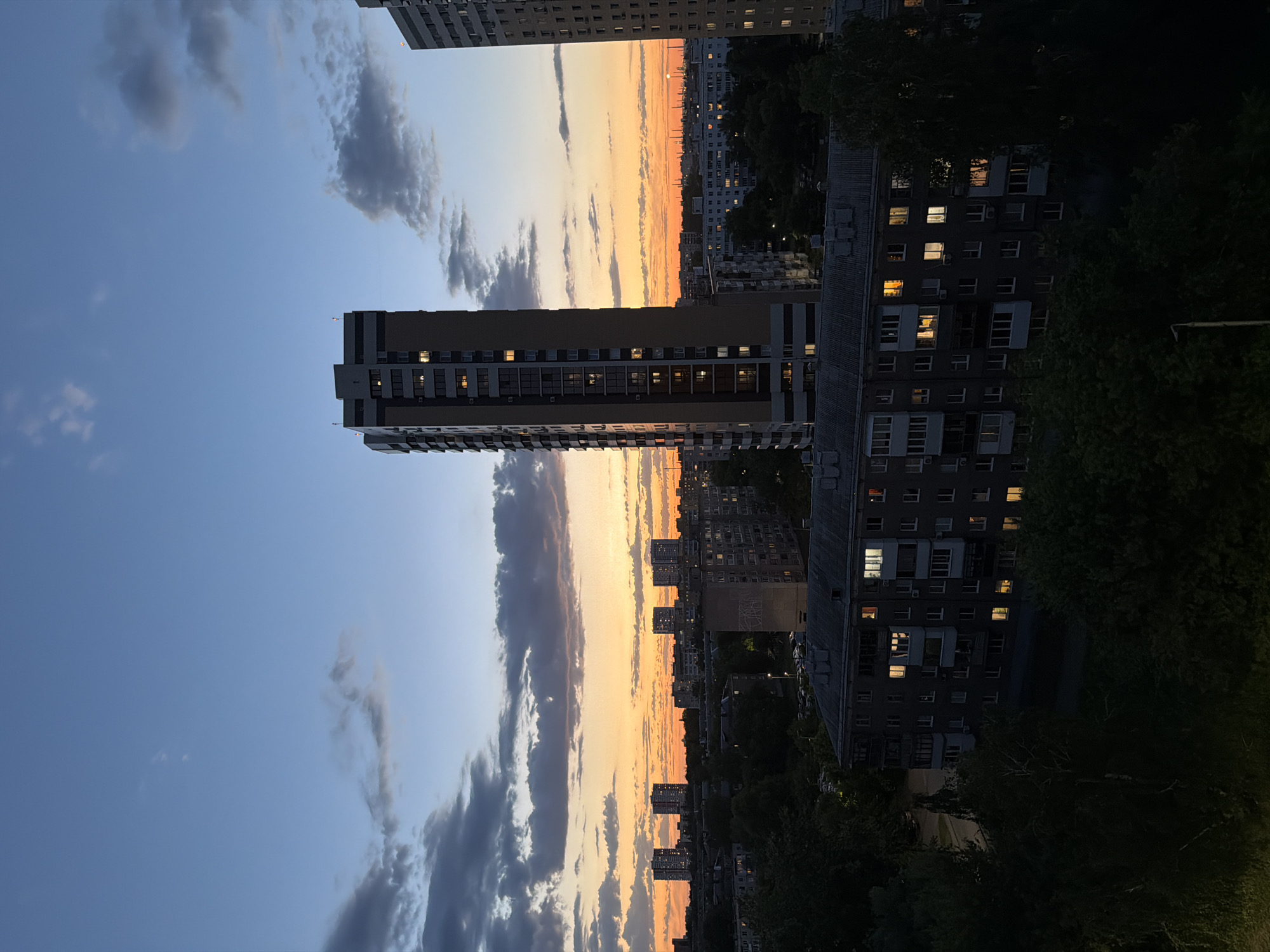 The image captures an urban scene at dusk, likely in the city of Unknown, Unknown. Several high-rise buildings are visible, some with illuminated windows, suggesting residential occupancy. The sky transitions from blue to orange and gray due to the sunset. Clouds are scattered across the sky. Green foliage indicates the presence of trees in the surrounding area.