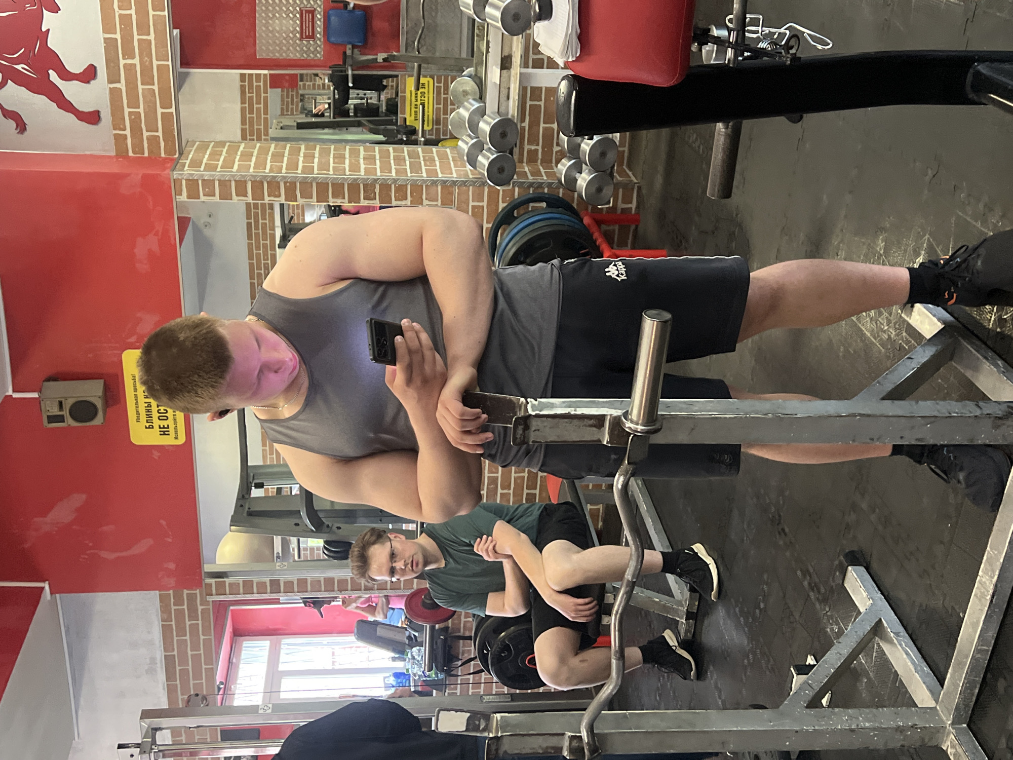 This image shows the interior of a gym, likely located in Saint Petersburg, Russia, based on the Russian language sign visible on the wall. The gym has brick walls and various exercise machines. A muscular man in a grey tank top and black shorts is standing in front of a weight machine, looking at his phone. Another man in glasses, wearing a green shirt and black shorts, is sitting on a different weight machine, looking forward. Weight plates and dumbbells are scattered around the room. The floor is covered with black mats.