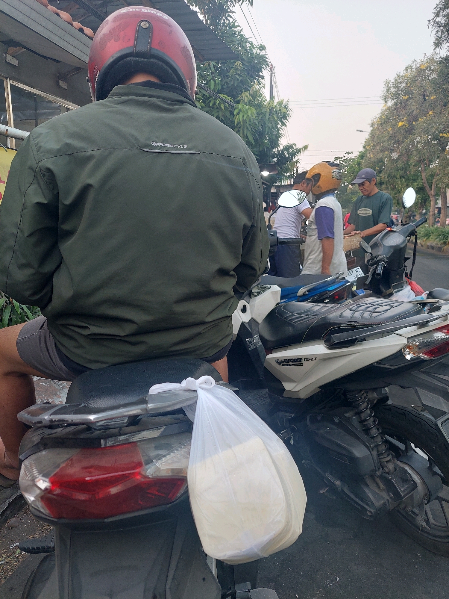 In Surabaya, Indonesia, the image captures a rear view of a person wearing a green jacket and red helmet, seated on a motorcycle. A white plastic bag containing what appears to be food is tied to the rear of the motorcycle. In the background, three other individuals are visible, two of whom are also on motorcycles. One person wears a yellow helmet and another a blue cap. The scene unfolds on a street lined with trees, suggesting an outdoor urban setting.