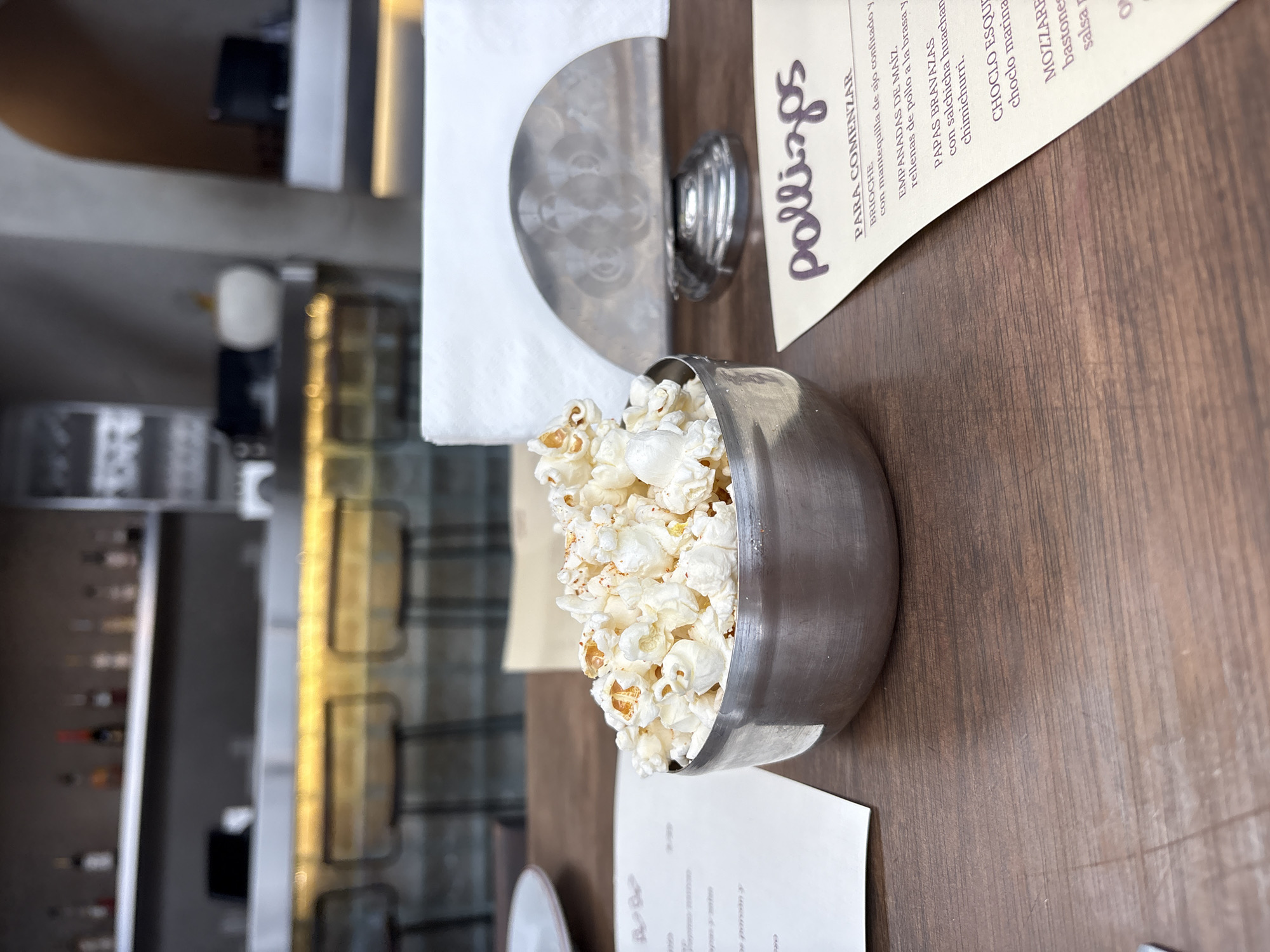 This image shows a close-up view of a metallic bowl filled with popcorn, placed on a wooden table. In the background, a blurred bar area is visible with bottles on shelves and stools. To the right of the popcorn, a menu with the word "pollizos" at the top is partially visible. The menu lists food items such as "BRIOCHE", "EMPANADAS DE MAÍZ", "PAPAS BRAVAS", and "CHOCLO ESQUEIX". The setting appears to be a restaurant in Miraflores, Peru, as suggested by the menu items. There are no people or discernible actions in the image.