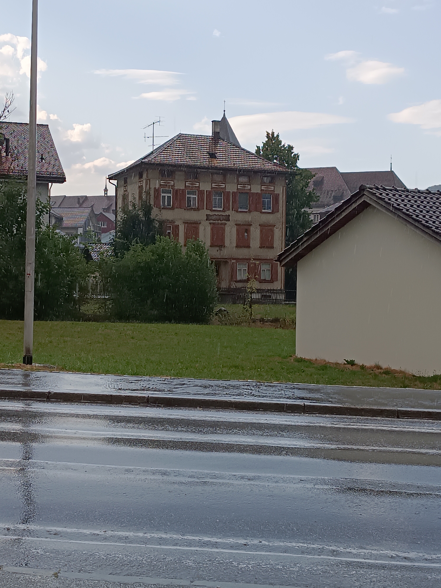 The image depicts a street scene in the city of Schwende-Rüte, Switzerland, during a rain shower. The foreground shows a wet asphalt road with visible reflections of the sky and surrounding objects. A concrete curb separates the road from a grassy area. In the middle ground, a well-maintained green lawn is present, with several trees and bushes. Beyond the lawn, a multi-story building with a tiled roof and shuttered windows is visible. The building's facade appears to be weathered. To the right, a portion of a lighter-colored building with a tiled roof is in view. In the background, several other buildings with tiled roofs and glimpses of a church spire can be seen. The sky is partly cloudy, with white clouds against a light blue background. Raindrops are visible throughout the image, indicating active precipitation. There are no people or animals visible in the scene. The overall impression is of a quiet, residential area experiencing a downpour.