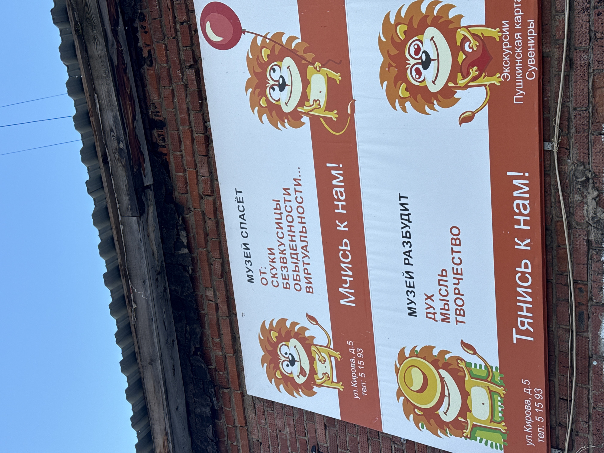 The image shows a sign in Russian promoting a museum, located on Kirov Street, building 5, phone number 5 15 93. The sign features cartoon lion figures and the phrases "Museum saves from boredom, tastelessness, ordinariness, virtuality..." and "Rush to us!" Another part of the sign says, "Museum awakens the spirit, thought, creativity" and "Reach out to us!" The sign also mentions excursions, the Pushkin card, and souvenirs. The sign is attached to a brick building under the edge of the roof. The sky is visible in the upper left corner.