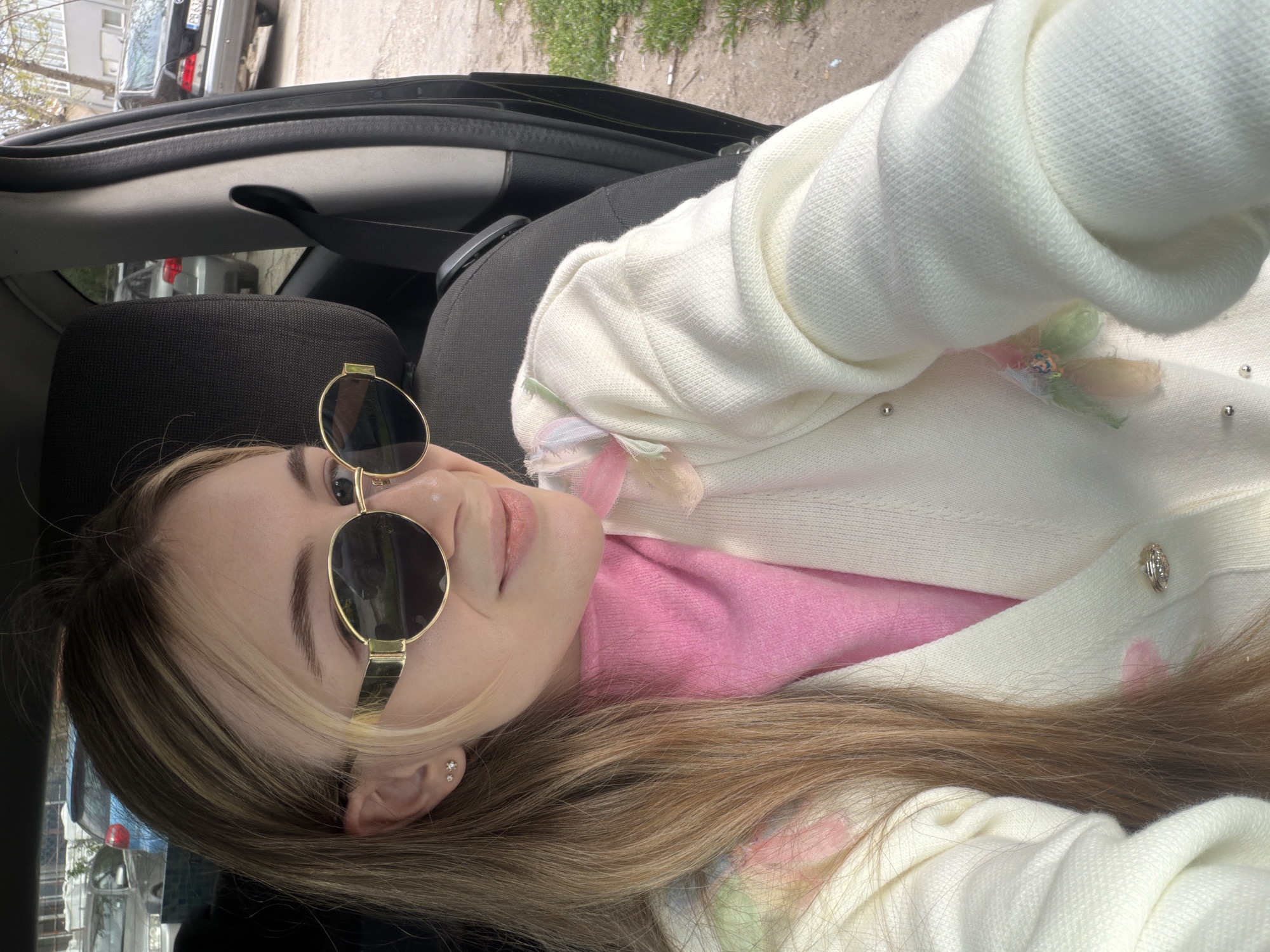 A woman with long, light brown hair is wearing sunglasses, a pink top, and a white jacket inside a car. The car is likely stationary. Outside the car, an urban environment is visible. The location is consistent with being in Plovdiv, Bulgaria, given the appearance of the vehicle in the background.