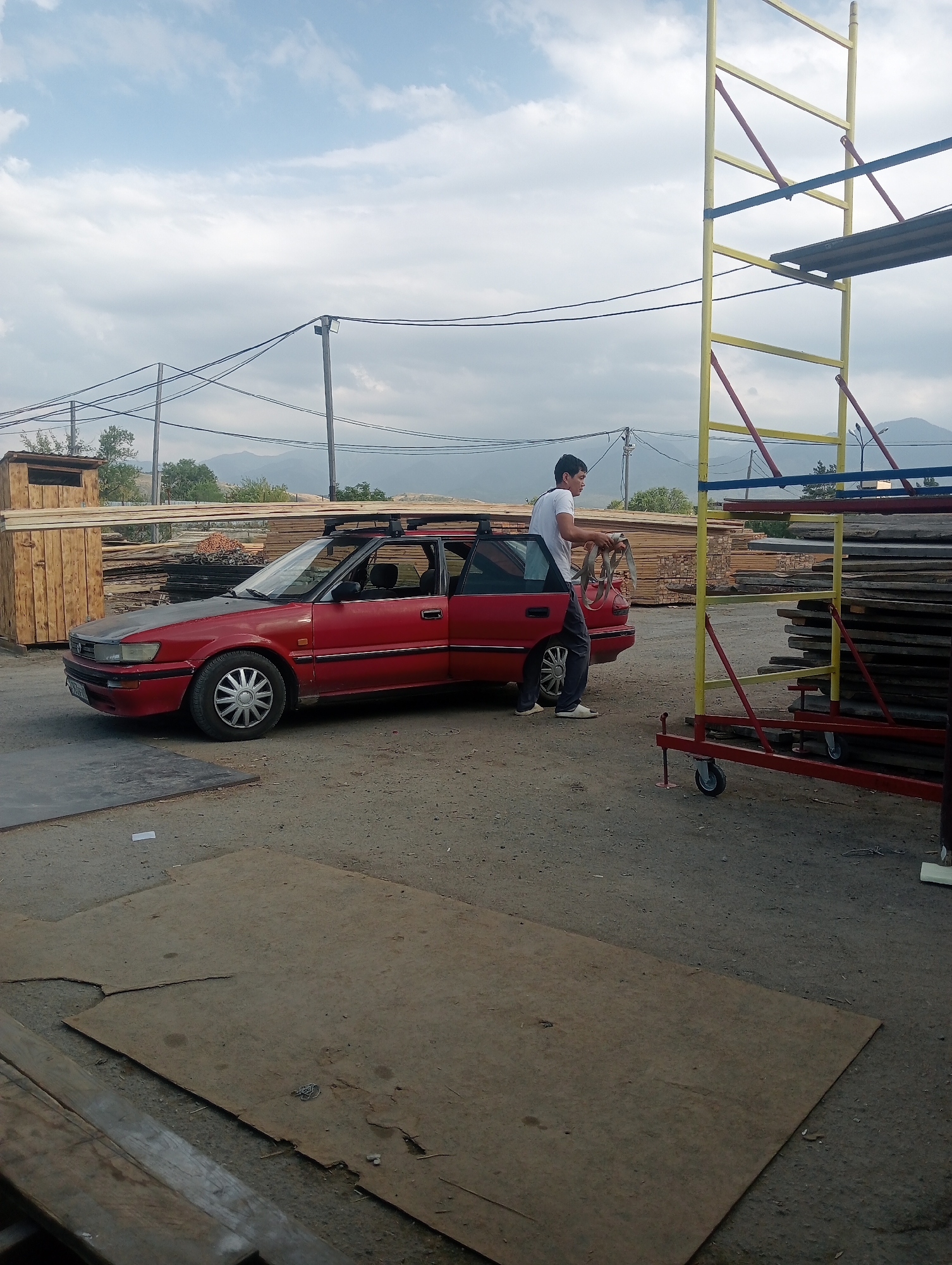 A man in a white t-shirt and dark pants is standing next to the open driver's side door of a red sedan. He is holding a coiled strap in his hands. The car has a roof rack and is parked on a dirt lot. In the background, there are stacks of lumber, scaffolding, and power lines. The scene appears to be an outdoor storage or construction area in Talgar, Kazakhstan. The sky is cloudy.