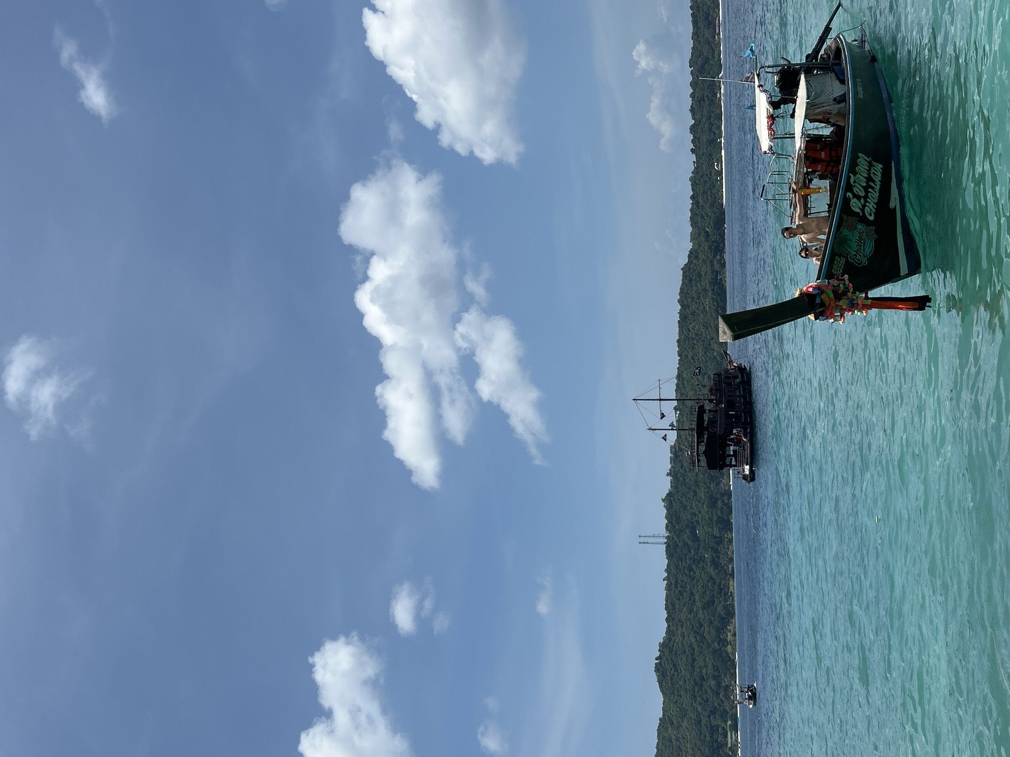 This is a horizontal, medium-shot, eye-level photograph of a tour boat in the ocean, with islands and a pirate-ship-style boat in the background. The water is turquoise and choppy, with light reflecting off the surface. In the foreground, a longtail boat, painted black with white and blue accents, is docked. Two individuals, a man and a woman, are standing on the boat. The man is shirtless and wearing swim trunks, while the woman is wearing a swimsuit. The woman is holding a life vest. The boat is decorated with colorful tassels and garlands. There is a thatched roof over the rear of the boat, and an outboard motor is visible. The name of the boat, "CHALLADA," is written in English and Thai on the side.
In the background, two larger boats are visible. The boat on the right is a black, pirate-ship-style vessel with two masts. The boat on the left is smaller and also dark in color, with a single mast. Behind the boats, a lush green island rises from the water, with buildings and communication towers visible on its slopes. The sky is a clear blue with scattered white clouds. The overall impression is a sunny day at sea, likely in a tourist destination known for its beautiful waters and boat tours.