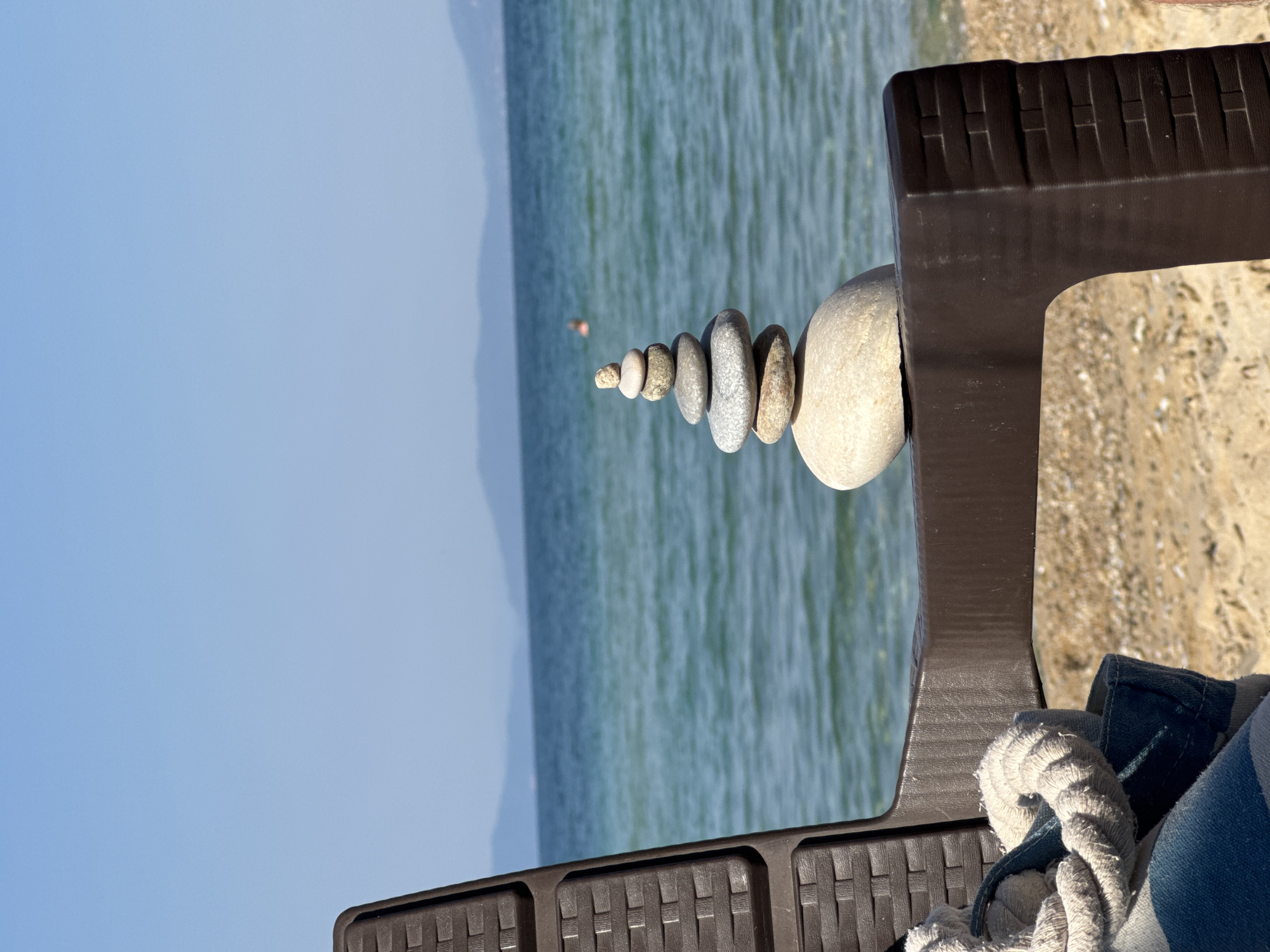 A stacked cairn of smooth, gray and beige stones rests on the armrest of a brown plastic beach chair on the sandy shore of Ofrynio Beach, Greece. The sea, a gradient of turquoise and deep blue, extends to the horizon, where hazy, mountainous islands are visible under a clear, pale blue sky. In the distance, a person is swimming in the ocean. A blue and white striped beach towel and a rope are draped over the armrest of the chair, partially obscuring the chair's textured surface. The sand beneath the chair is a light tan color and scattered with small, white shells. The image is captured from a low angle, with the beach chair prominently in the foreground, framing the seascape.