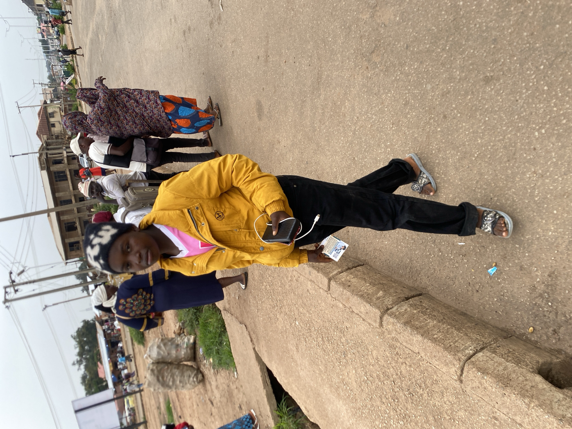 A young person wearing a yellow jacket, black pants, and sandals walks on a paved road. They are holding a black device and a small paper in their right hand. In the background, several other people are visible, along with buildings and two large bags of what appears to be produce. The scene is set in a street in Bukuru, Nigeria, indicated by the presence of a street sign with "Bukuru" visible. The weather appears overcast.