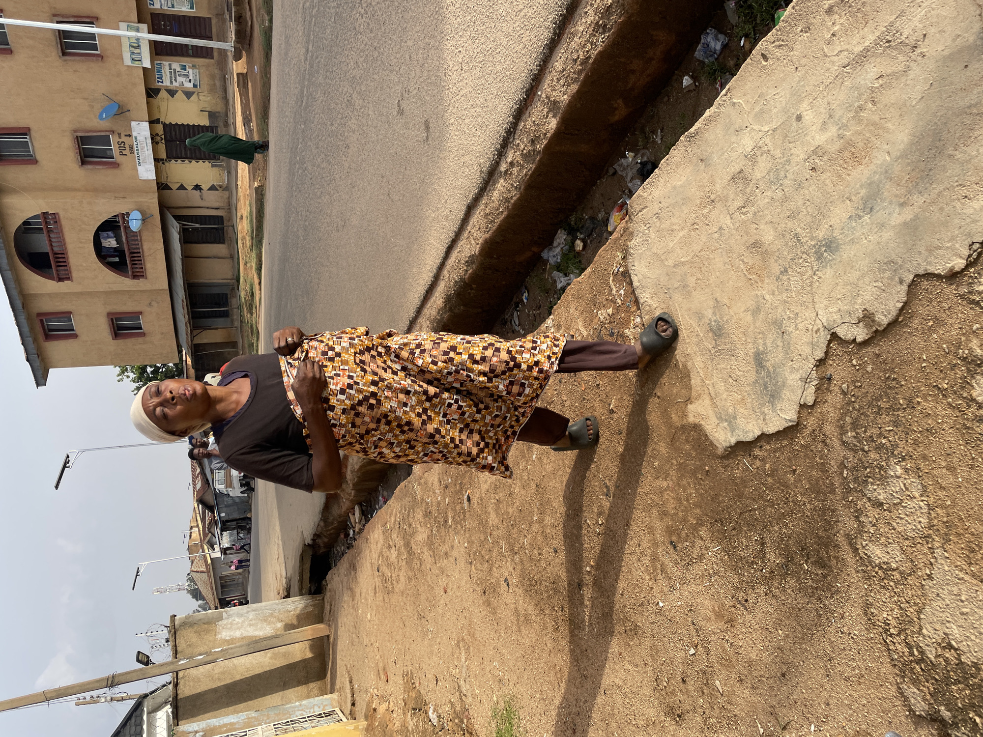 An elderly woman in a dark-colored shirt, patterned skirt, and white head covering is walking on a cracked concrete sidewalk next to a street in Jos, Nigeria. The street has a deep drainage ditch running along its edge filled with debris. Buildings line the opposite side of the street. Power lines and light posts are visible in the background.