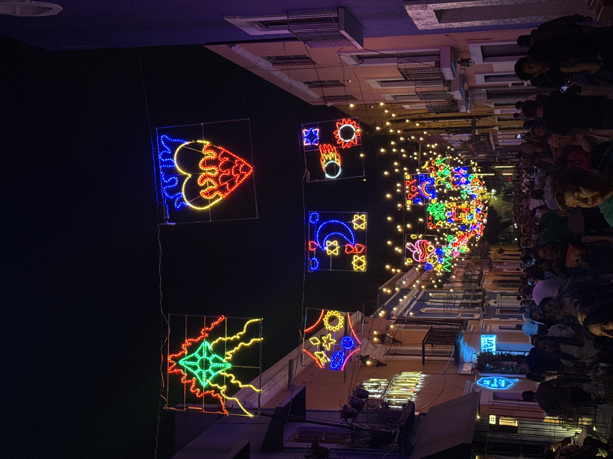 The image shows a nighttime street scene in Puebla, Mexico, decorated for a festival. The street is lined with buildings, and the upper stories are visible on both sides. The buildings are mostly lit, with some having signs and decorations.
Crowds of people are visible on the right side of the image, packed along the street. They appear to be watching a procession or display.
Hanging from cables above the street are several large, illuminated decorations. These are stylized images with bright, colorful lights. The decorations include abstract designs, geometric shapes, and representations of celestial bodies.
String lights extend down the center of the street, creating a glowing canopy effect. Metal scaffolding and stage equipment are visible along the lower portion of the buildings.
