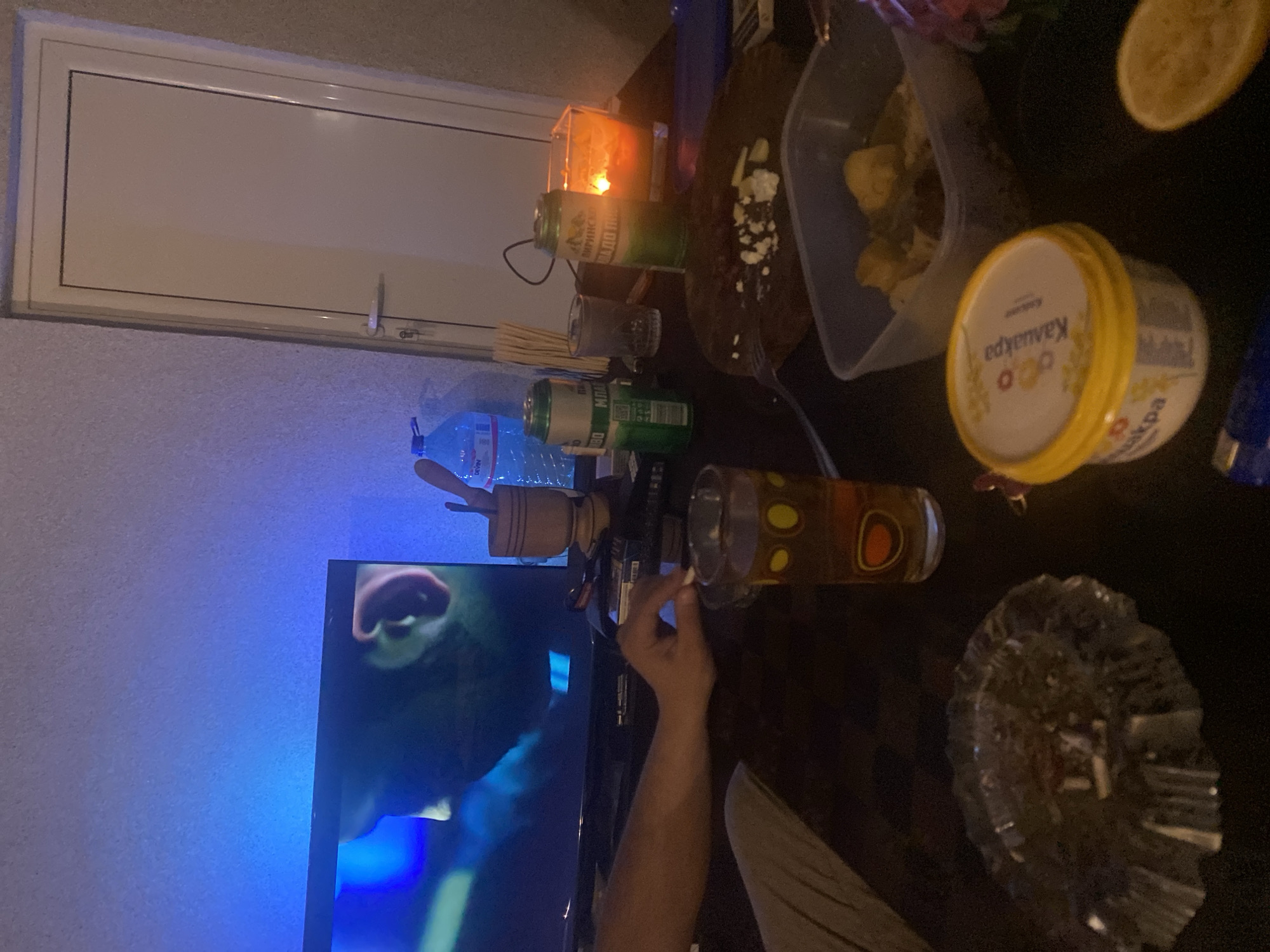 This image captures an indoor, dimly lit evening scene, likely a casual gathering or an individual relaxing. The perspective is from above a table, which is cluttered with various items. A person's arm and hand are visible in the lower right, seemingly reaching for something or holding a small item near a patterned glass containing a brown liquid, likely beer. An ash tray filled with cigarette butts and ash sits to the right, suggesting smoking.
On the table, several beverages and snacks are arranged. There's a blue plastic bottle labeled "DEVIN" (water), two green cans (one identifiable as "Пиринско" beer, another with "МОР"), and a yellow-lidded container marked "Кауфланд" (likely a dairy product like spread cheese or yogurt). A clear plastic container holds what appears to be fried potatoes or other snack food, accompanied by a fork. Half a lemon slice is also on the table. A lit candle in a glass holder provides warm illumination, contrasting with the cool blue light emanating from a television screen on the left, which displays a close-up of a person's face or head.
Other items include a wooden mortar and pestle, a small clear glass, and a black remote control. A white-framed window with a light-colored blind or panel, displaying "LED" on its handle, is visible in the background. The visible text on the items, such as "Пиринско" and "Кауфланд," are consistent with the provided location context of Belopoptsi, Bulgaria. The overall atmosphere suggests a relaxed, low-key indoor activity.