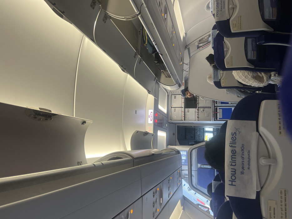 The image displays the interior of an IndiGo airplane cabin, captured in a vertical (portrait) orientation, looking down an aisle from an aisle seat towards the front or middle of the aircraft. The cabin is brightly illuminated by warm white overhead lights.
In the immediate foreground, the back of a passenger's head with dark hair is visible, seated in an aisle seat. Further down the aisle, other passengers are seated, and a person, possibly a flight attendant or passenger, is partially visible near the galley area. The seats are upholstered in blue, featuring white headrest covers.
Overhead bins line both sides of the cabin; some are open, revealing luggage inside, while others are closed. An "EXIT" sign in red lettering with a directional arrow is prominently visible on the left side of the cabin. The aircraft's galley or service area, with various compartments and service carts, can be seen in the distance.
Visible text on a headrest cover in the foreground clearly states: "How time flies", "18 years of IndiGo", and "#IndiabyIndiGo", identifying the airline. Another partially visible sign on a seat back in the lower right corner reads "FASTEN SEAT BELT WHILE SEATED" and includes some text in Hindi. The scene suggests passengers are either boarding, deplaning, or the aircraft is in flight. Given the location context of Guwahati, India, this is an IndiGo flight to or from Guwahati.