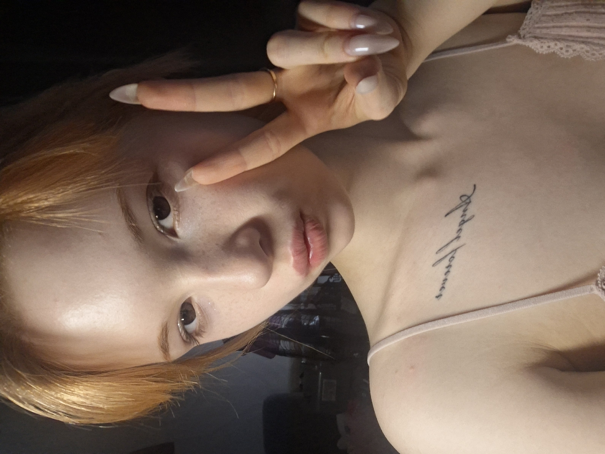 This close-up portrait, oriented diagonally, features a young individual with fair skin and light reddish-blonde hair. The person is making a peace sign with their right hand, held up near their face, and is looking directly at the camera.
Notable features include dark brown eyes, a scattering of light freckles across the nose and cheeks, and slightly parted lips with a natural pink, glossy finish. The individual's fingernails are long, almond-shaped, and appear to be artificial, with a translucent nude or light pink color. A simple gold ring is visible on the right ring finger.
On the upper right side of the chest, a small cursive tattoo runs vertically. The text, deciphered from the image, reads "Stay forever". The subject is wearing a light pink or nude-colored top or bra with a delicate lace trim on the strap. The background is dark and out of focus, suggesting an indoor setting with low or artificial light. There are no specific visual cues to indicate the location as Chaikovsky, Russia, or the time of day or weather conditions. The overall mood is casual and intimate.