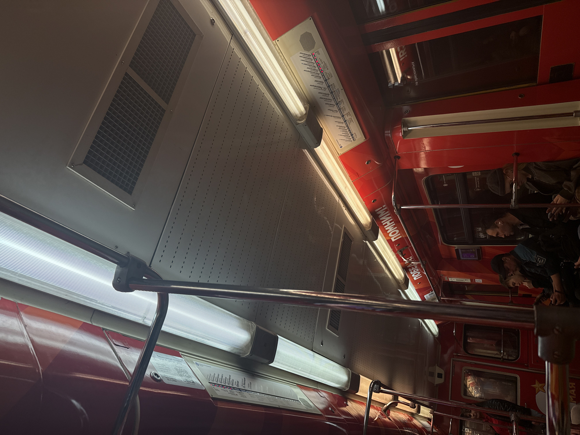 Here is a description of the image:
The image depicts the interior of a Moscow Metro train, likely the Arbatsko-Pokrovskaya Line based on the route map visible. The interior features typical train elements like overhead lighting, ventilation panels, handrails, and windows. There are passengers present within the train. The image captures the subway line route information with station names in Cyrillic script. There are signs or labels, notably "ПОБЕДА ПОМНИМ!" in Cyrillic, and a warning "Не прислоняться" (Do not lean).
