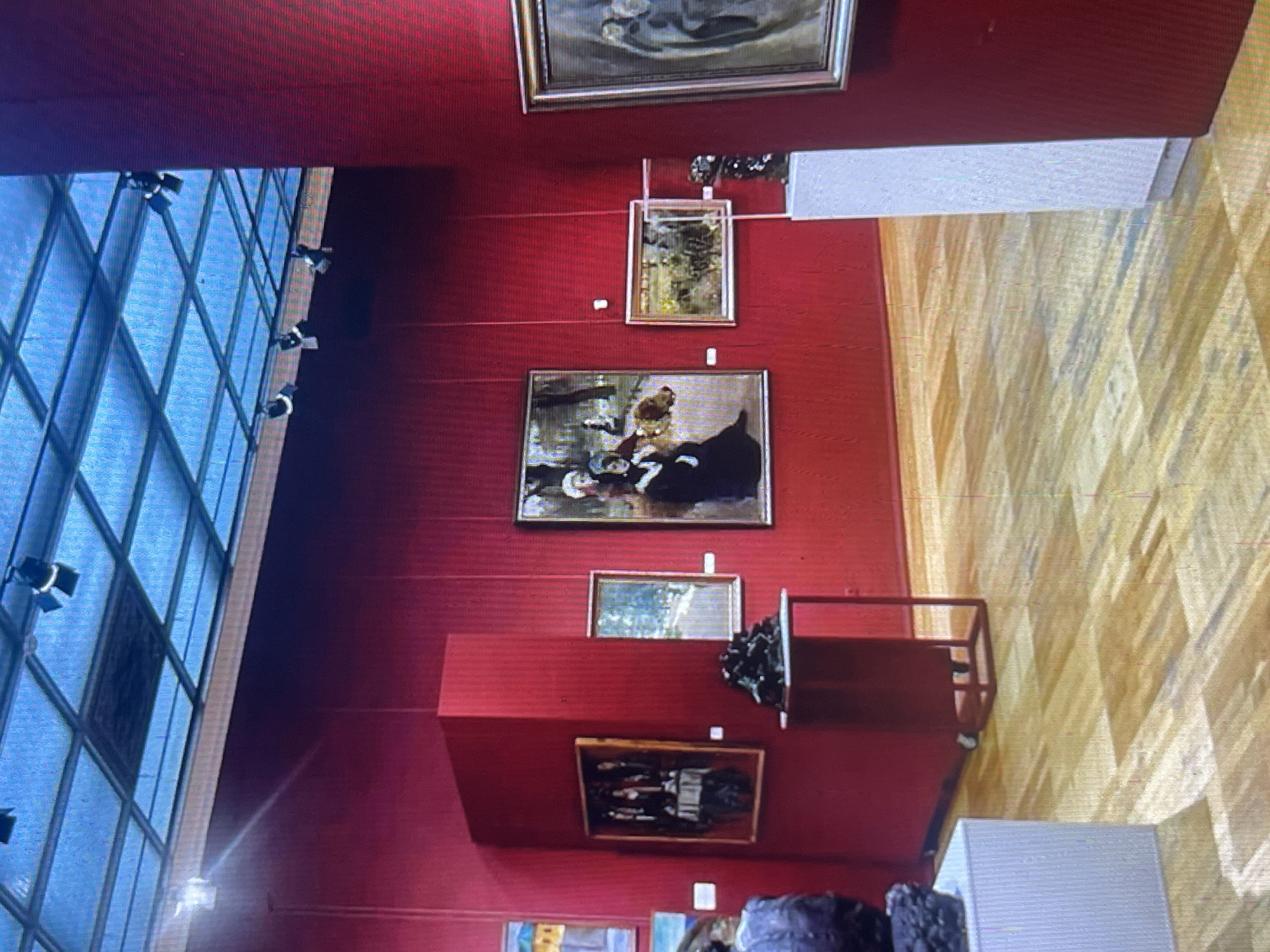 The image shows an interior view of an art gallery with red walls and a wooden floor. Several paintings are displayed on the walls, with frames of varying sizes and colors. In the foreground, a white pedestal displays a dark sculpture, and to its left, another white pedestal shows a portion of a dark sculpture. To the right of the first pedestal, a red display stand with a metal frame holds another dark sculpture. A glass-enclosed display is visible on the floor in front of a painting. The ceiling features a blue grid structure with spotlights. The image is a still photograph taken from a medium-low angle. The location is not specified as Bishkek City, Kyrgyzstan.