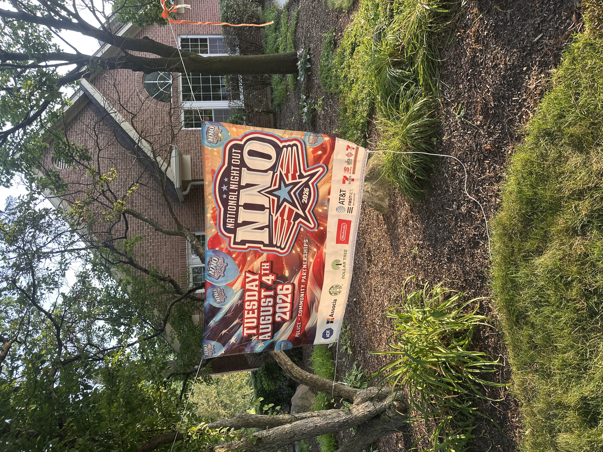 The image shows a banner hanging between two trees in front of a house. The banner advertises "National Night Out" and displays "Tuesday August 4th 2026". It also features the logos of various sponsors, including ADT, Associa, Starbucks, Dollar Tree, Nintendo, AT&T, 7-Eleven, and Taco Bell. The house appears to be a brick residence with white-trimmed windows and a large arched window above the entrance. The scene is set outdoors, with green grass and shrubs in the foreground, and sunlight casting shadows. The location is identified as Schaumburg, United States of America.