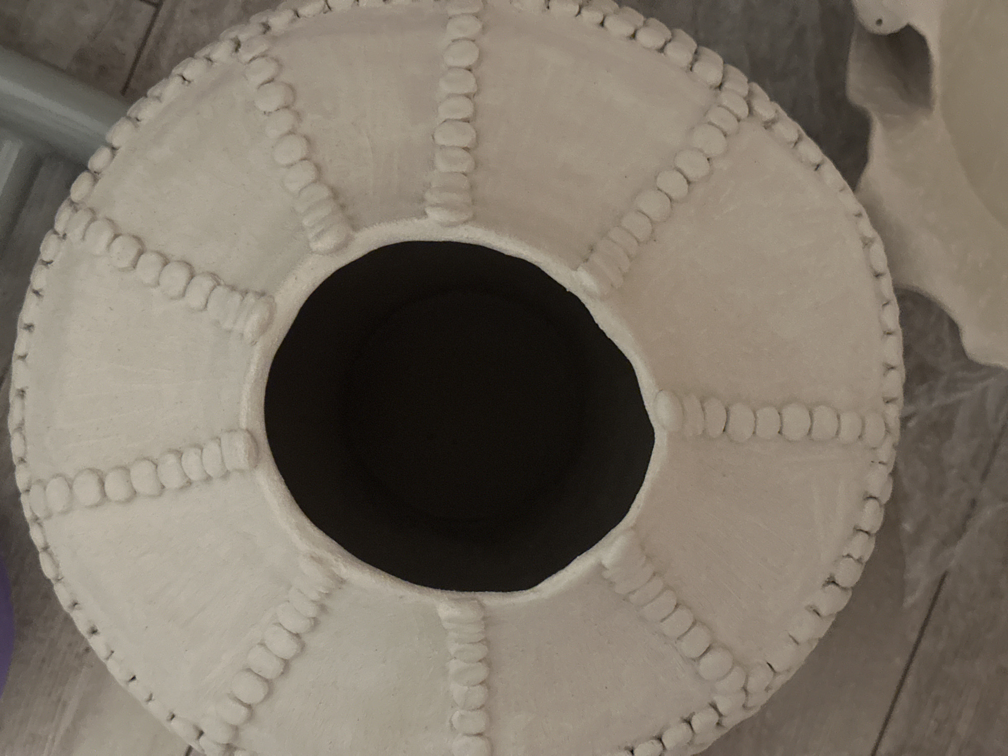 The image shows a top-down view of a beige ceramic vase with a circular opening. The vase is decorated with raised, bead-like patterns arranged in radial lines and a circular border. The surface of the vase appears to be textured and slightly rough. The opening of the vase is dark and appears to be empty. The vase is placed on a surface that resembles wooden floorboards. There are no people or animals visible in the image. The environment suggests an indoor setting, possibly a home or studio. No specific location in Nevinnomyssk, Russia can be identified from the image.