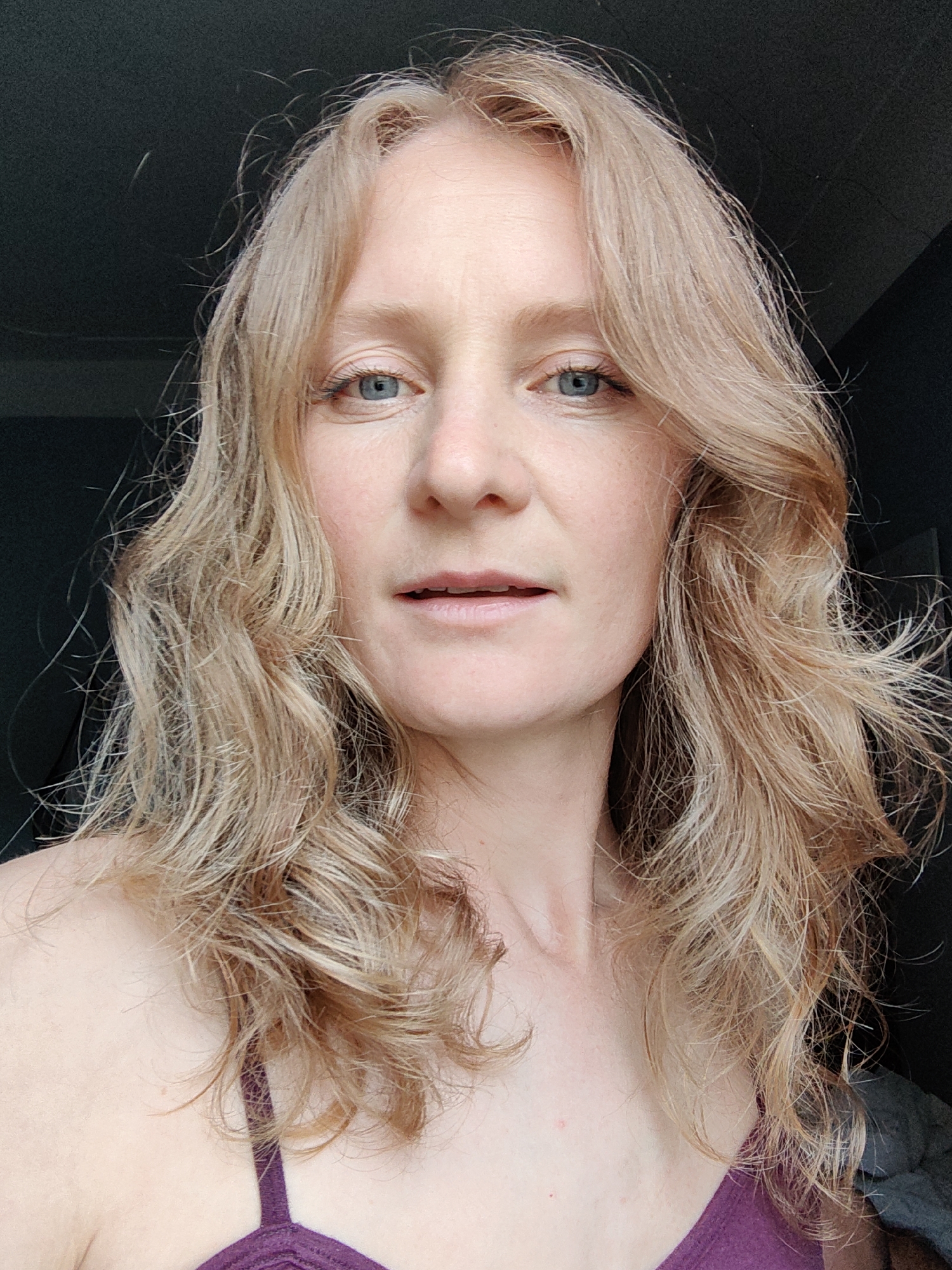 A close-up, medium shot shows a fair-skinned woman with light blonde wavy hair and blue eyes. She is wearing a purple top. The background is out of focus and dark. The setting's location is not discernable from the provided image.