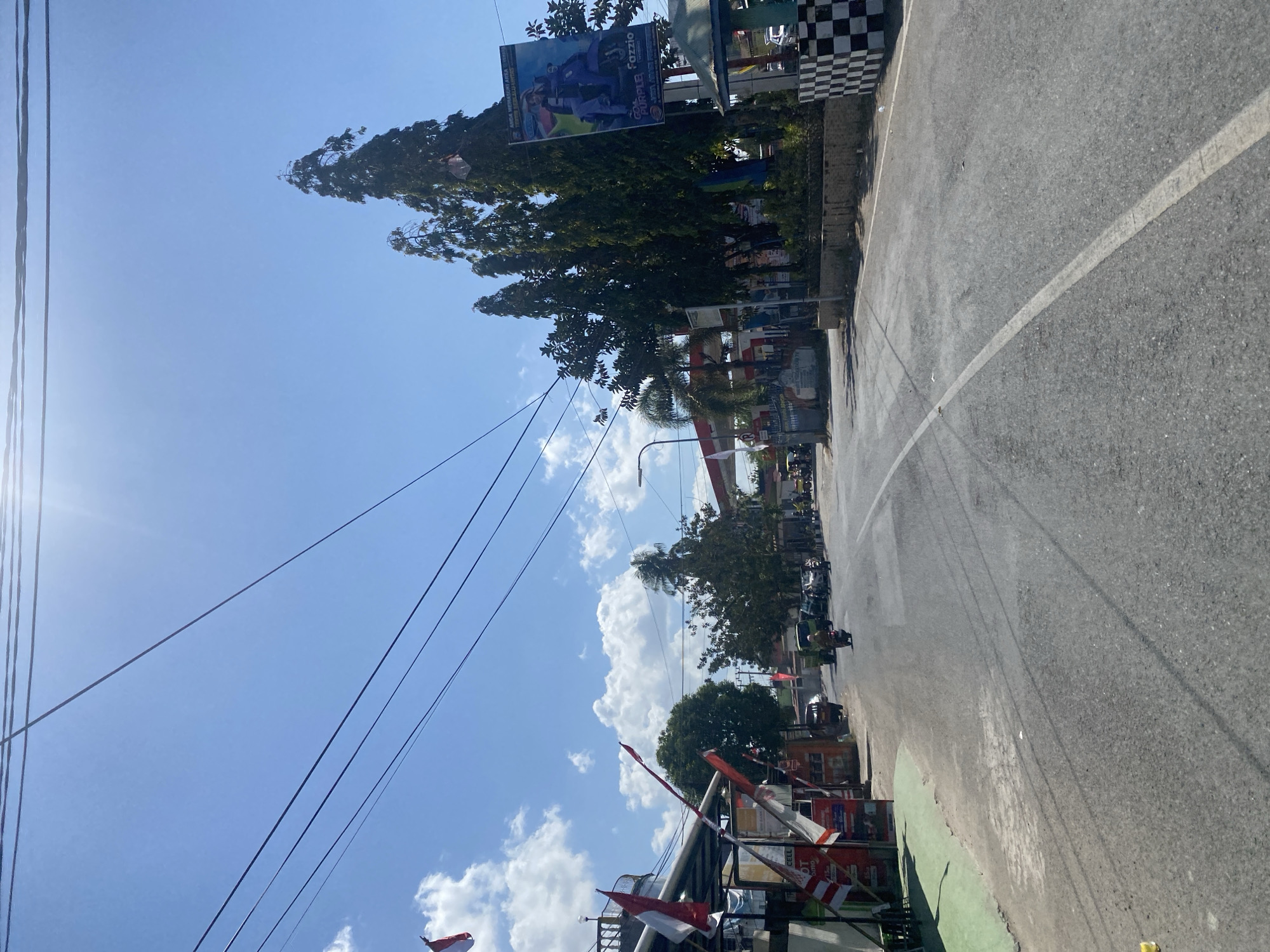 The image depicts a street scene in Bulukumba, Indonesia, on a sunny day with a clear blue sky and scattered clouds. The road, a paved asphalt surface with a white dividing line, curves to the right. On the left side of the road, there are buildings with awnings decorated with Indonesian flags. Various shops and stalls are visible, with some displaying advertisements. A few people are seen walking and riding motorcycles.
On the right side of the road, a gas station with red and white branding is prominent. Tall trees line the area behind the gas station. A large billboard advertising "Fazzio" and featuring images of a scooter and a person is also visible. A checkered black and white barrier marks the edge of a property. Several vehicles, including cars and motorcycles, are present on the road, suggesting moderate traffic. Power lines crisscross the sky above the scene. The overall impression is of a typical roadside environment in a Southeast Asian city.
