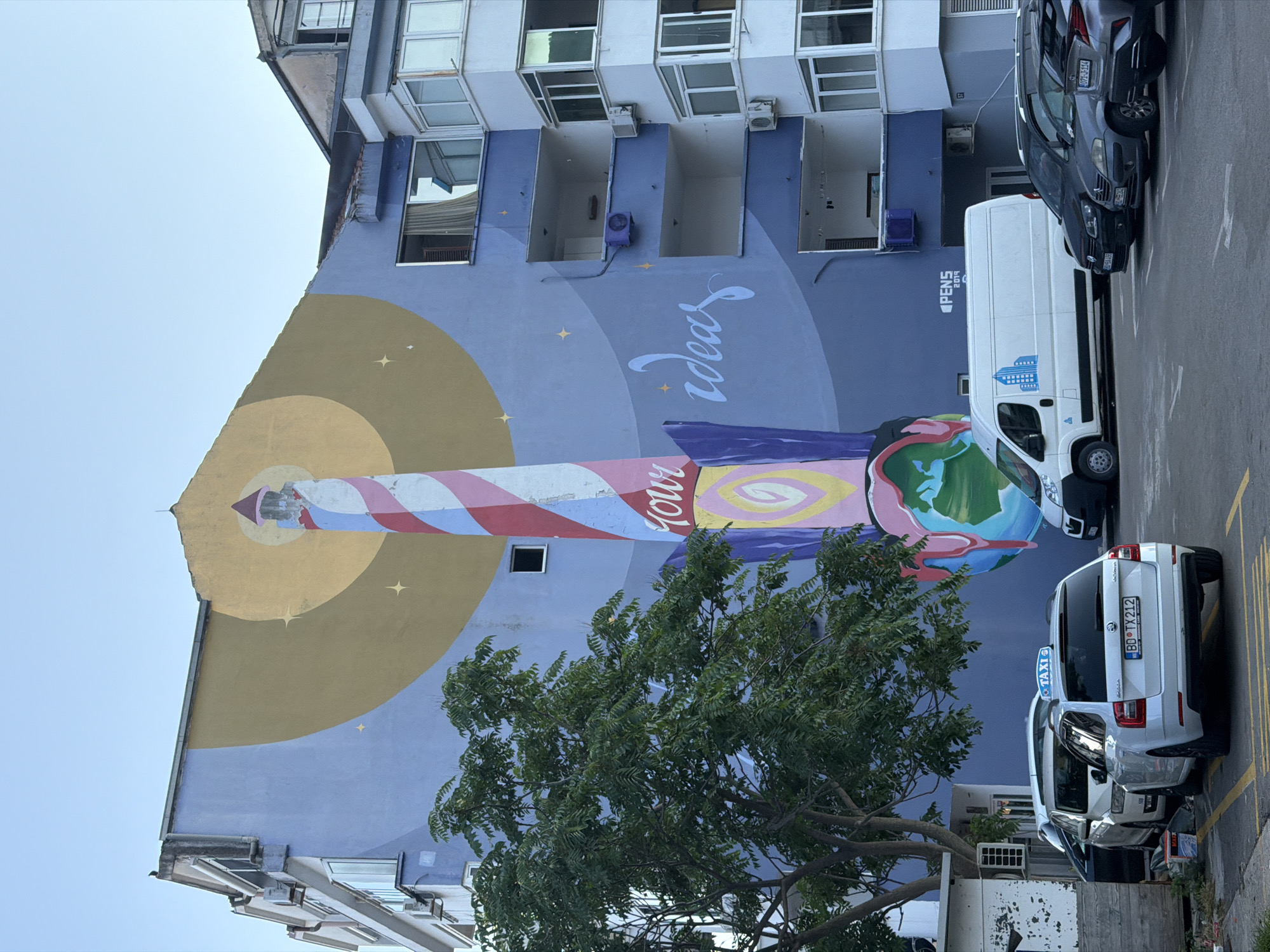 This is a photograph of a building in Budva, Montenegro, featuring a large mural. The mural depicts a stylized lighthouse or rocket, painted in vibrant colors like red, white, and blue, against a soft blue and yellow background. The lighthouse/rocket is adorned with imagery of a globe at its tip, suggesting a theme of exploration or dreams. The word "ideas" is artistically written on the wall in a flowing script, also in white.
The building itself appears to be an apartment complex, with multiple windows and balconies visible. Some windows are open, revealing glimpses of the interior, and air conditioning units are mounted on the exterior walls. The overall aesthetic of the building suggests it may be older, with some areas showing signs of wear.
In the foreground and to the right, a parking lot is visible, with several cars parked. Among them are a white van and a white taxi, the latter clearly marked with "TAXI" and displaying a license plate that reads "BD TX212". The presence of parked vehicles suggests it's daytime and a normal day in the city. A green tree with lush foliage partially obscures the mural and the building. The sky is clear and bright, indicating fair weather. The photograph is taken from a slightly low angle, looking up at the building and mural. The style of the mural and the general setting evoke a sense of urban art and a coastal city atmosphere. The text "PENS 2019" is visible on the building near the white van.