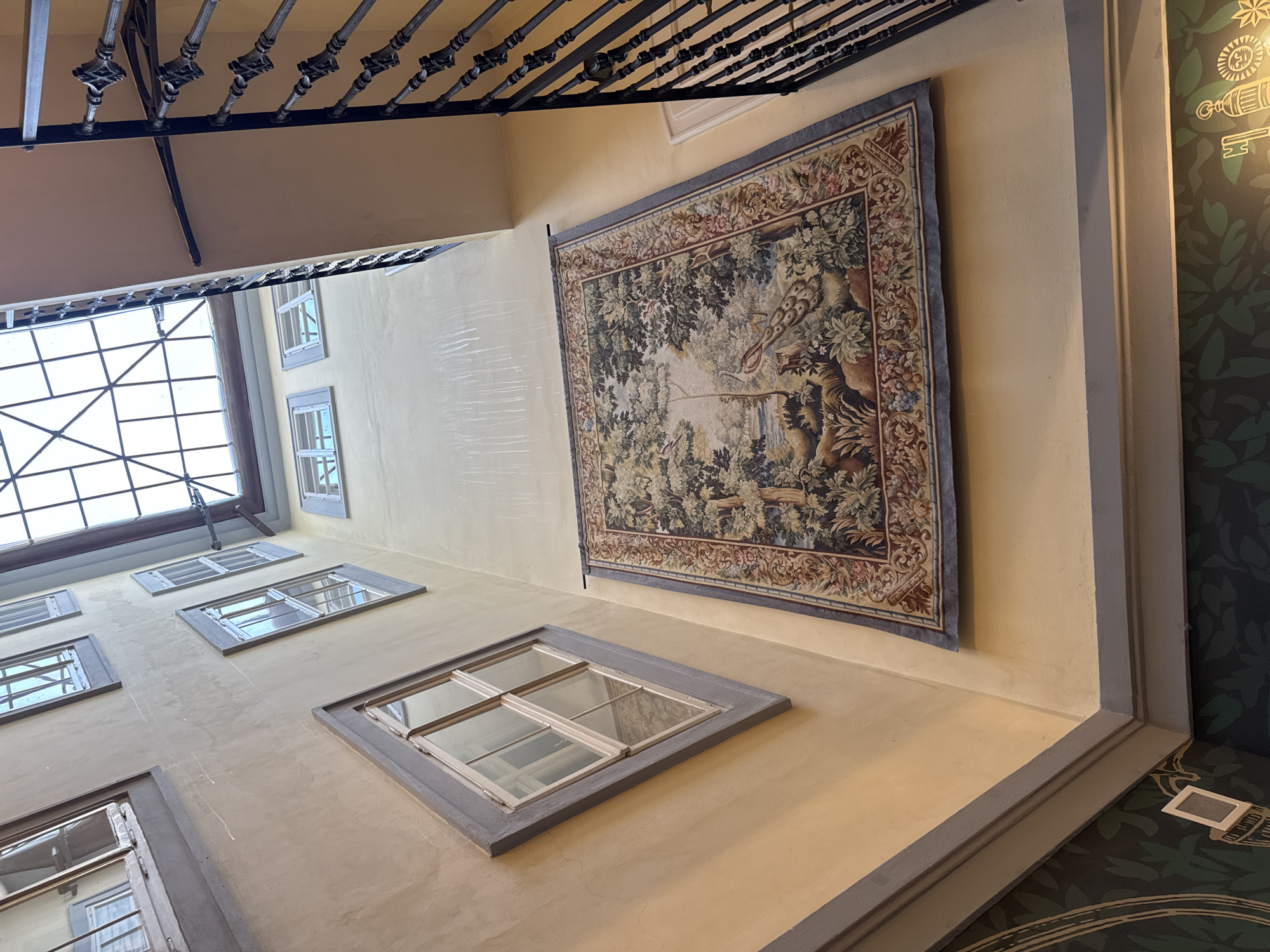 An interior view looks up at a skylight in a building in Prague, Czechia. The image captures an atrium with a yellow-painted wall, several windows, and a large tapestry hanging on the wall. A dark metal railing of a staircase is visible on the right side of the image. The tapestry depicts a pastoral scene with trees, a bird, and a body of water. The skylight above is a grid of glass panes. The walls show some faint vertical water stains. The bottom of the image shows a portion of dark green wallpaper with a floral pattern.