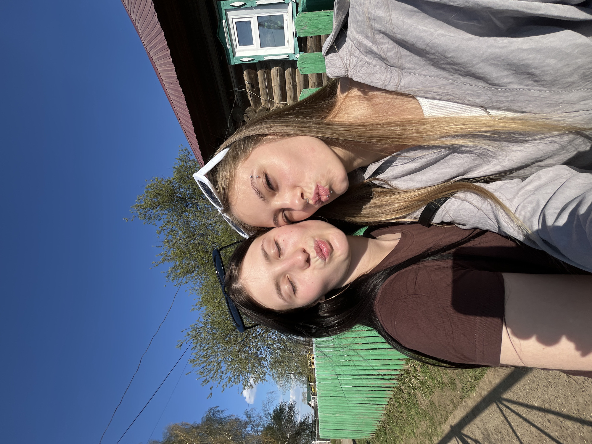 Two young women are posing for a selfie outdoors in Фоки, Russia. The woman at the top has long blonde hair and is wearing white sunglasses, a gray hoodie, and a white top. She has eyebrow piercings and a lip ring. The woman at the bottom has dark hair and is wearing dark sunglasses and a brown shirt. Both women have their eyes closed and are puckering their lips. Behind them, there is a green wooden building with a small window, a tree, a blue sky, and a red roof. The area appears to be somewhat rural.