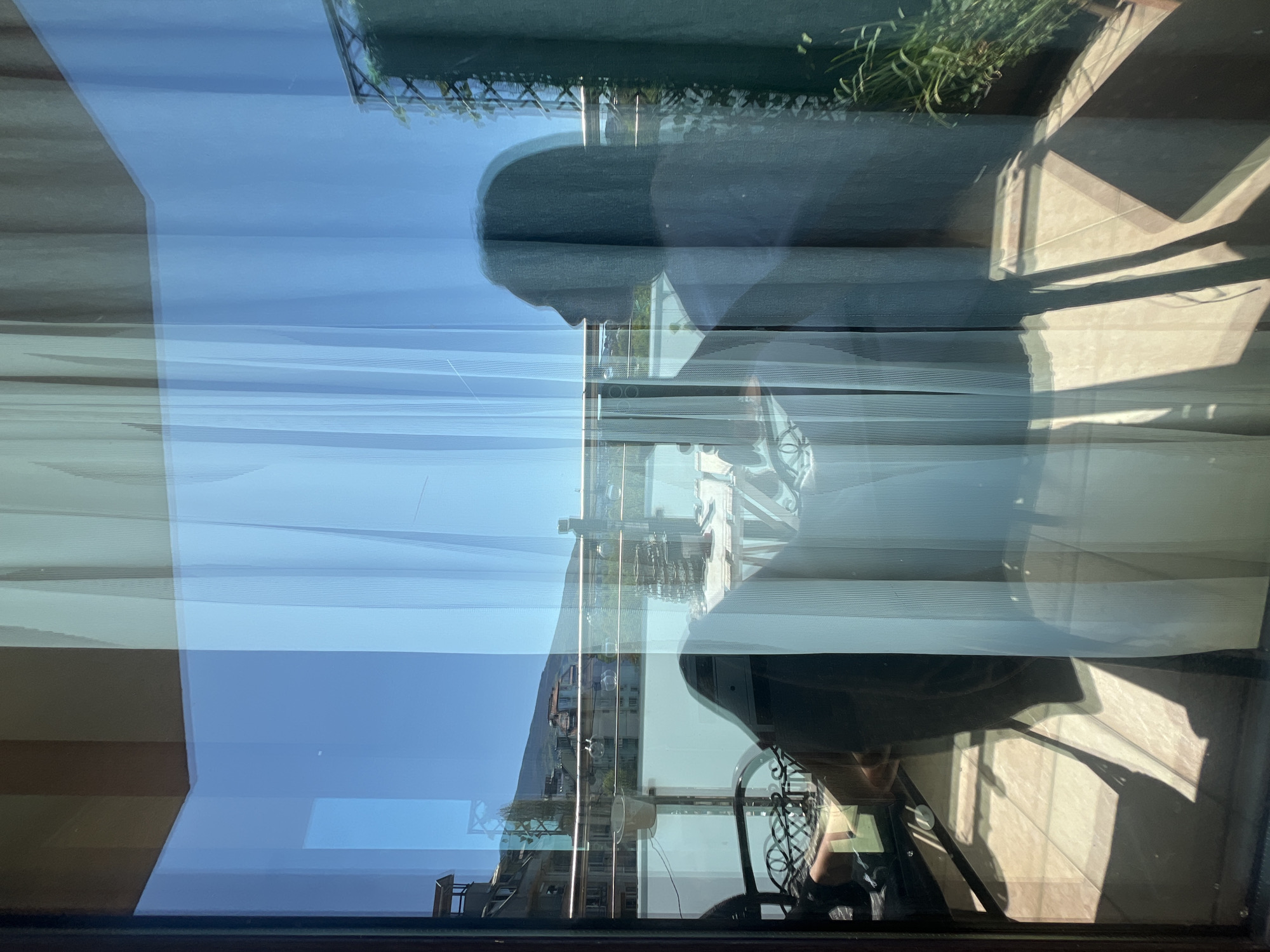 The image shows a reflection in a window. The reflection reveals parts of an interior, including a chair and possibly a person sitting in it, a balcony or patio with some vegetation, and the outside world. The reflection shows what seems to be a building, some greenery (possibly a mountain in the background), and clear blue sky. The location can not be identified, but the image suggests it is in the city of Sofia, Bulgaria.