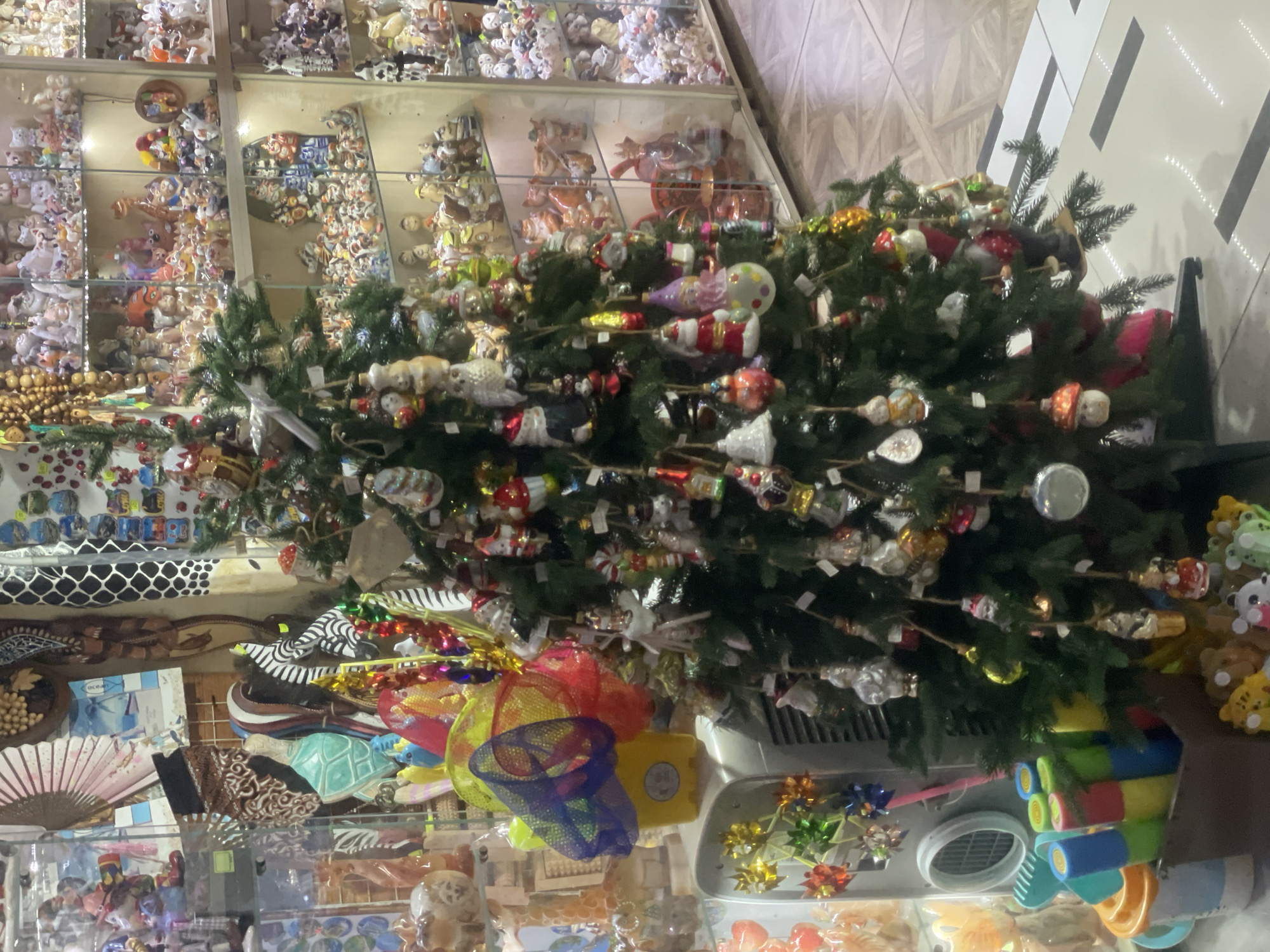 A decorated Christmas tree stands in what appears to be a retail establishment, possibly a souvenir shop or market, in Sochi, Russia. The tree is adorned with numerous small ornaments, including figures resembling Santa Claus, bells, animals, and baubles. The background displays shelves filled with a variety of ceramic figurines, trinkets, and decorative items, consistent with a shop setting. To the left of the tree, a yellow bucket with a colorful net is visible, along with what looks like a ventilation unit and various children's toys. The flooring is tiled with a pattern of white and dark gray rectangles. The overall scene depicts a festive, cluttered retail environment.