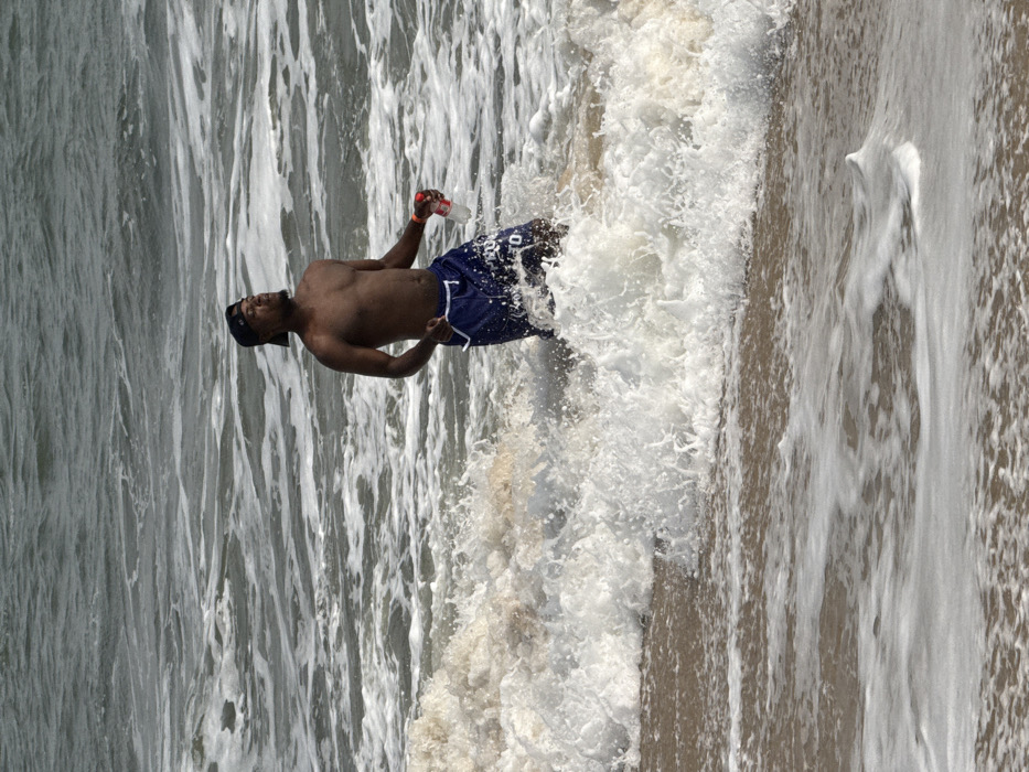 An adult male stands waist-deep in the ocean at Mopo Onijebu, Nigeria. The subject is shirtless, displaying a dark complexion, and wears dark blue swim trunks along with a dark baseball cap worn backward. He holds a clear plastic bottle with a red cap in his right hand and has a red band on his right wrist. White ocean waves are actively breaking around his midsection, generating substantial foam and spray. His head is tilted slightly upward, and his eyes appear closed or squinting. The background consists of turbulent, dark greyish-green ocean water, while the foreground shows wet sand with a layer of receding foamy water.