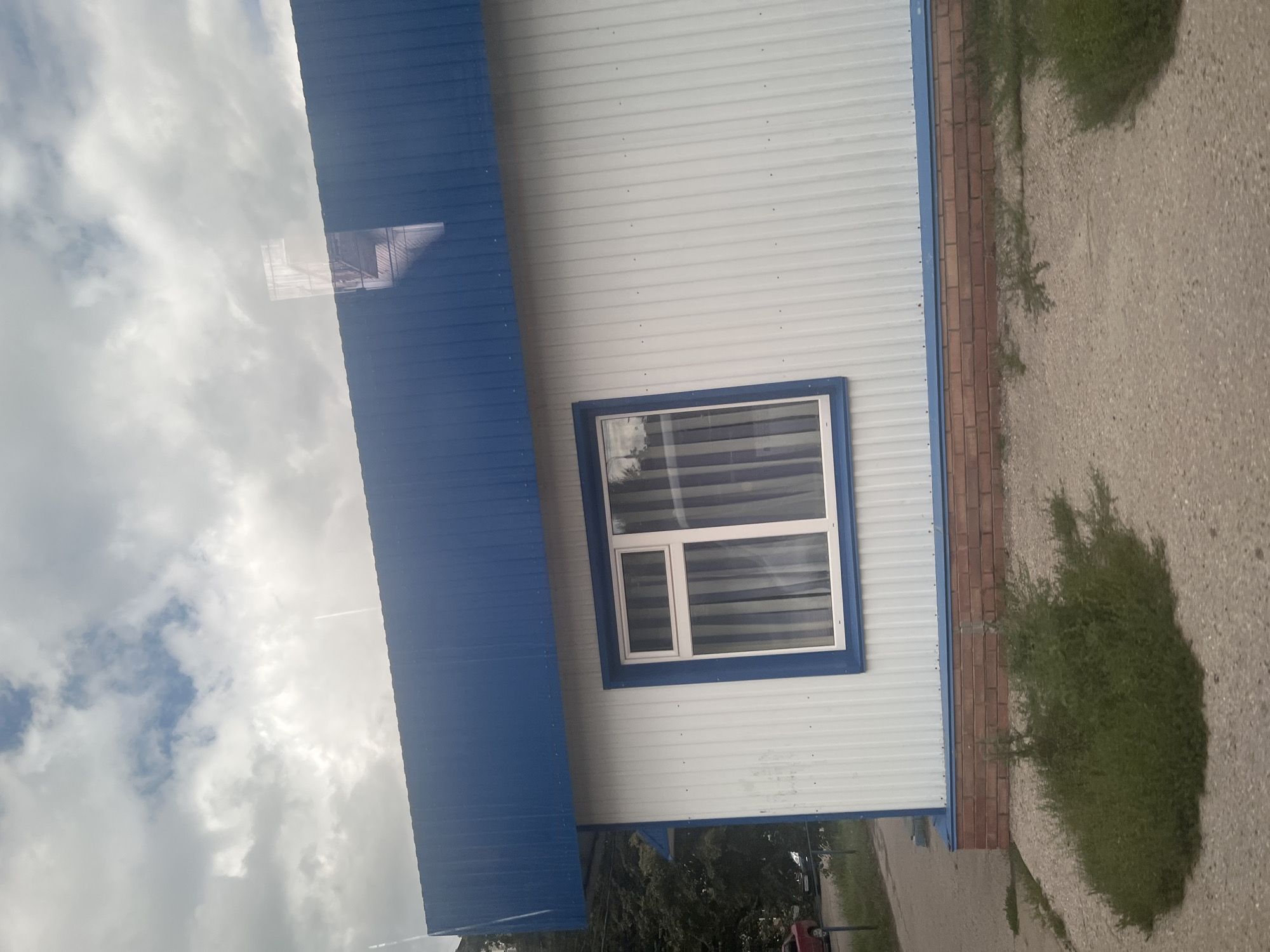 A side view of a small, white, corrugated metal building with a blue metal roof is presented. A single white-framed window with vertical blue and white striped curtains is visible on the building's facade. The building is situated on a gravelly surface with patches of green grass and weeds growing around it. The foundation of the building appears to be constructed with red brick. In the background, trees and a hint of a road with vehicles can be seen. The sky above is cloudy with patches of blue. The scene is likely located in Ulyanovsk, Russia, based on the provided context.