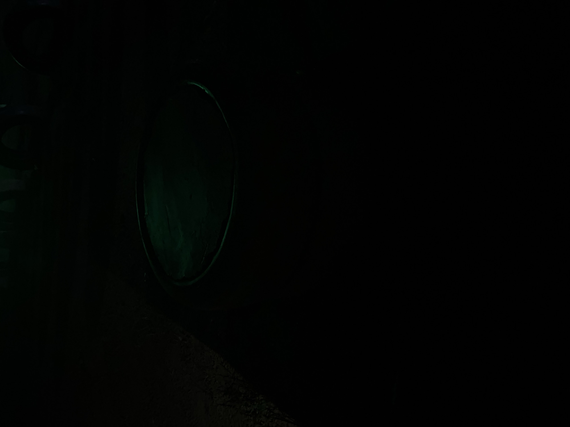 The image shows a dimly lit outdoor scene, with the majority of the frame in darkness. In the upper right portion of the image, a circular object with a reflective surface is visible, illuminated by a green light. The object appears to be a shallow basin or a lid. The surrounding environment is indistinct due to the low light conditions, but appears to be ground or soil. There are no people or discernible activity present in the scene. The location is specified as Howayi, Nigeria.