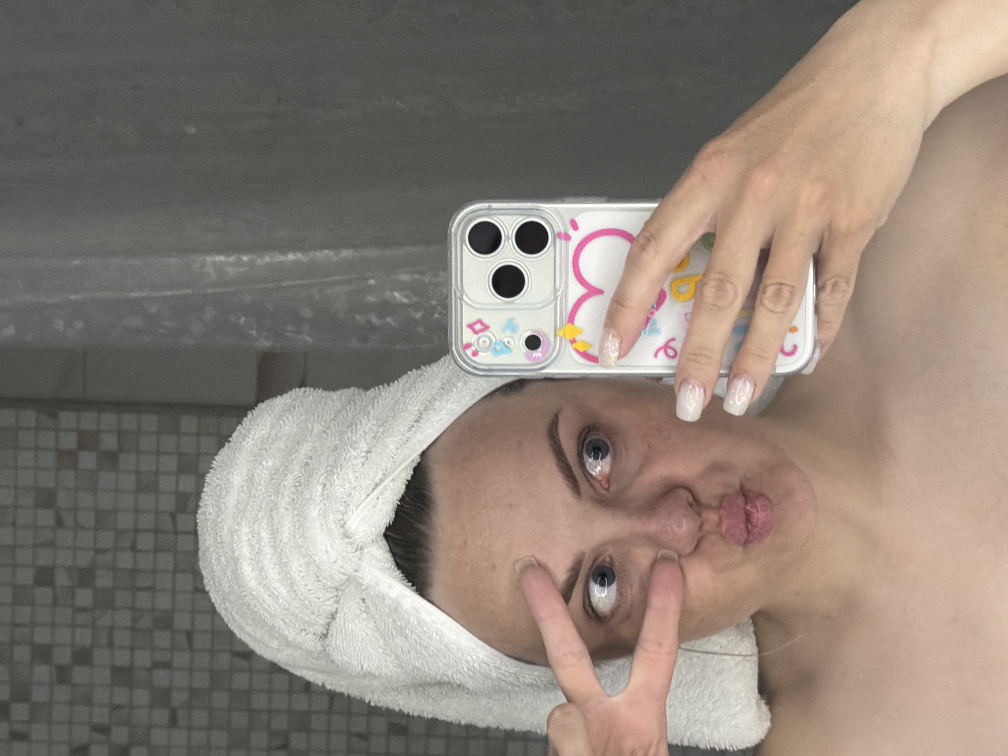 A woman poses for a selfie in what appears to be a bathroom. She is wearing a white towel wrapped around her head. She holds a phone in her right hand, the phone has a clear case with a cartoon design. She makes a peace sign with her left hand near her face, with pursed lips. The background includes tiled walls. The location of this image is unknown, there is no verifiable location within the city of Votkinsk, Russia.