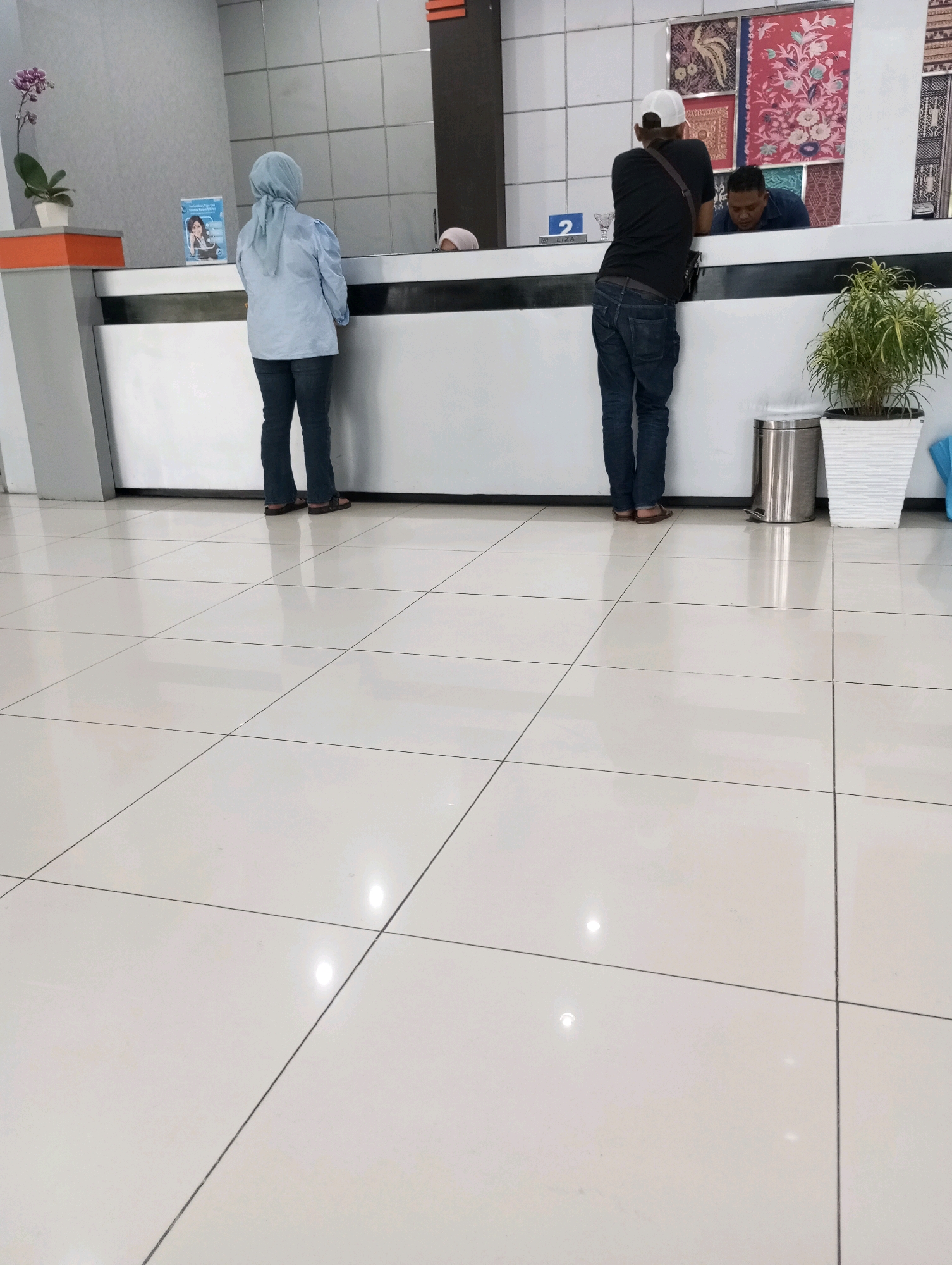 The image shows a counter area, likely within a government office or bank in Padang Panjang, Indonesia. A woman wearing a light blue hijab and a long-sleeved shirt stands at the counter, facing away from the camera. To her right, a man wearing a black t-shirt, blue jeans, and a white baseball cap leans over the counter, interacting with someone out of frame. Another man, wearing a blue shirt, is partially visible behind the counter, seemingly attending to the man in the black t-shirt. The counter itself is white with a dark stripe along the top. Behind the counter, decorative textiles with floral and bird motifs are displayed on the wall. A potted plant with green leaves sits to the right of the counter, next to a silver trash can. The floor is tiled with large, light-colored square tiles. The environment appears clean and orderly.