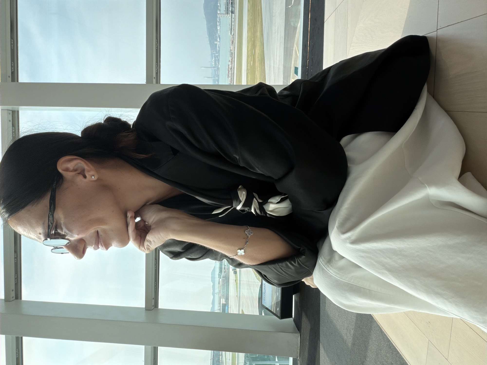 A medium shot shows a woman with dark hair pulled back into a bun, wearing eyeglasses, a black blazer, and loose white pants. She is sitting near a large window with a view of an airport tarmac and harbor. The woman is looking to her left with her chin resting on her hand. A clover-shaped bracelet is visible on her wrist. The background shows the expansive window overlooking an airport with planes and a distant industrial area with a body of water and hills. The floor is tiled and wooden. The lighting suggests it is daytime.