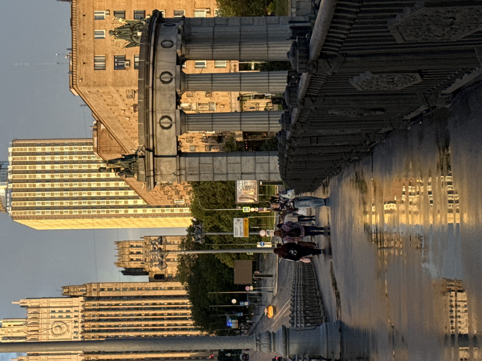 This is an eye-level shot from a bridge in Moscow, Russia, featuring a walkway with several people strolling across it. The bridge is lined with an ornate, dark metal railing on the right. The wet surface of the walkway reflects the golden hues of sunlight hitting large buildings in the background.
Prominently featured on the bridge is a stone rotunda-like structure with large columns and decorative elements. Beyond this, the Moscow skyline is visible with several distinct buildings. To the left, a tall, imposing building with vertical accents and a clock face is partially obscured by a large lamppost. To its right, a narrower, ornate historical building with battlements stands. Further to the right, a very tall, slender building with a grid of windows is bathed in golden light. Behind these, a series of orange brick buildings with numerous windows are visible.
Several people are walking on the bridge. On the left side of the walkway, four individuals are visible, two walking away from the camera and two walking towards it. On the right side of the walkway, a single person is walking towards the camera. The overall atmosphere is one of a late afternoon or early evening with warm, directional lighting. The presence of reflections suggests recent rain.