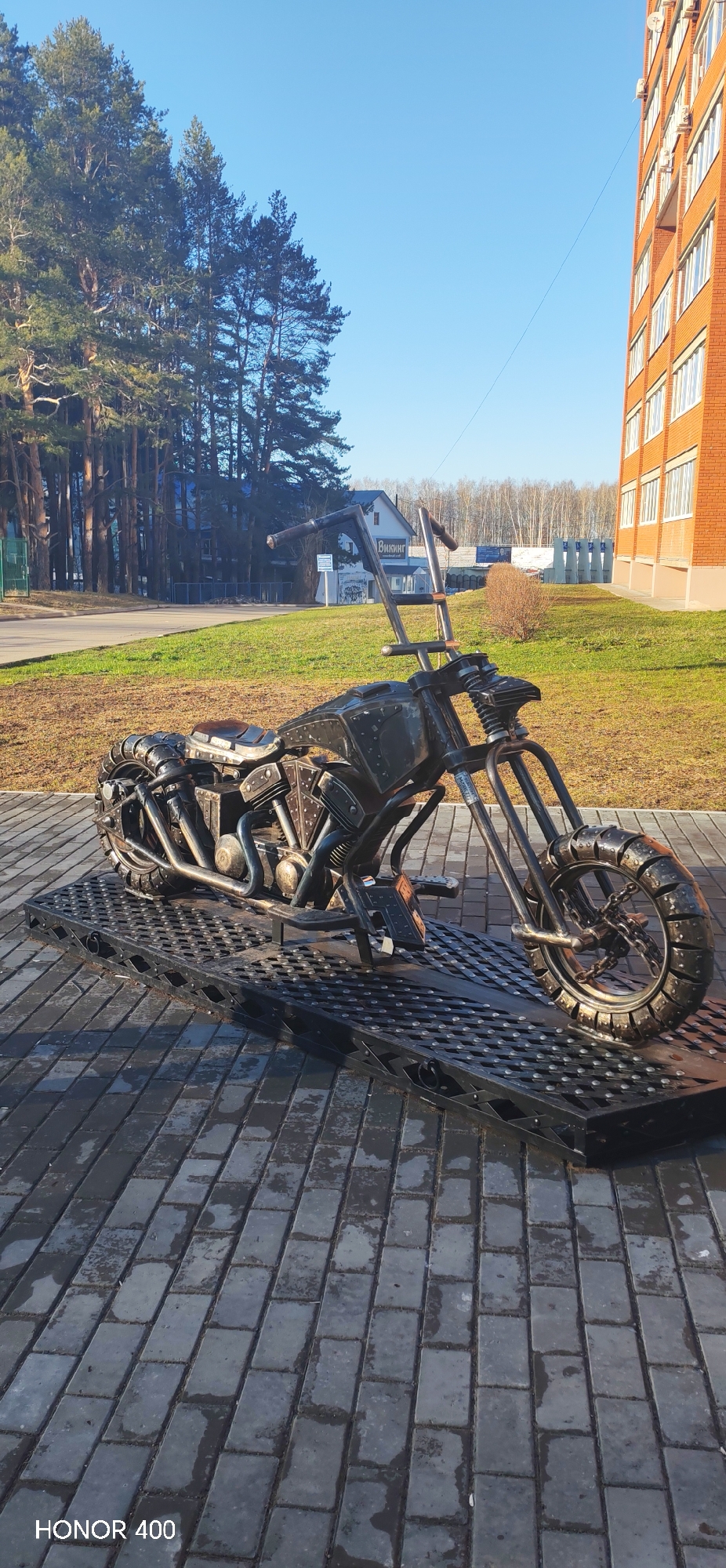 The image depicts a metal sculpture of a motorcycle situated on a black metal platform, placed on a paved walkway. Behind the sculpture, there's a building with a sign in Russian that says "Викинг", partially obscured. Further in the background, there are trees and additional buildings. To the right of the sculpture, there's a red brick building. The sky is blue, suggesting a clear day. The photo was taken in the city of Chaikovsky, Russia. The sculpture is the primary subject of the image.