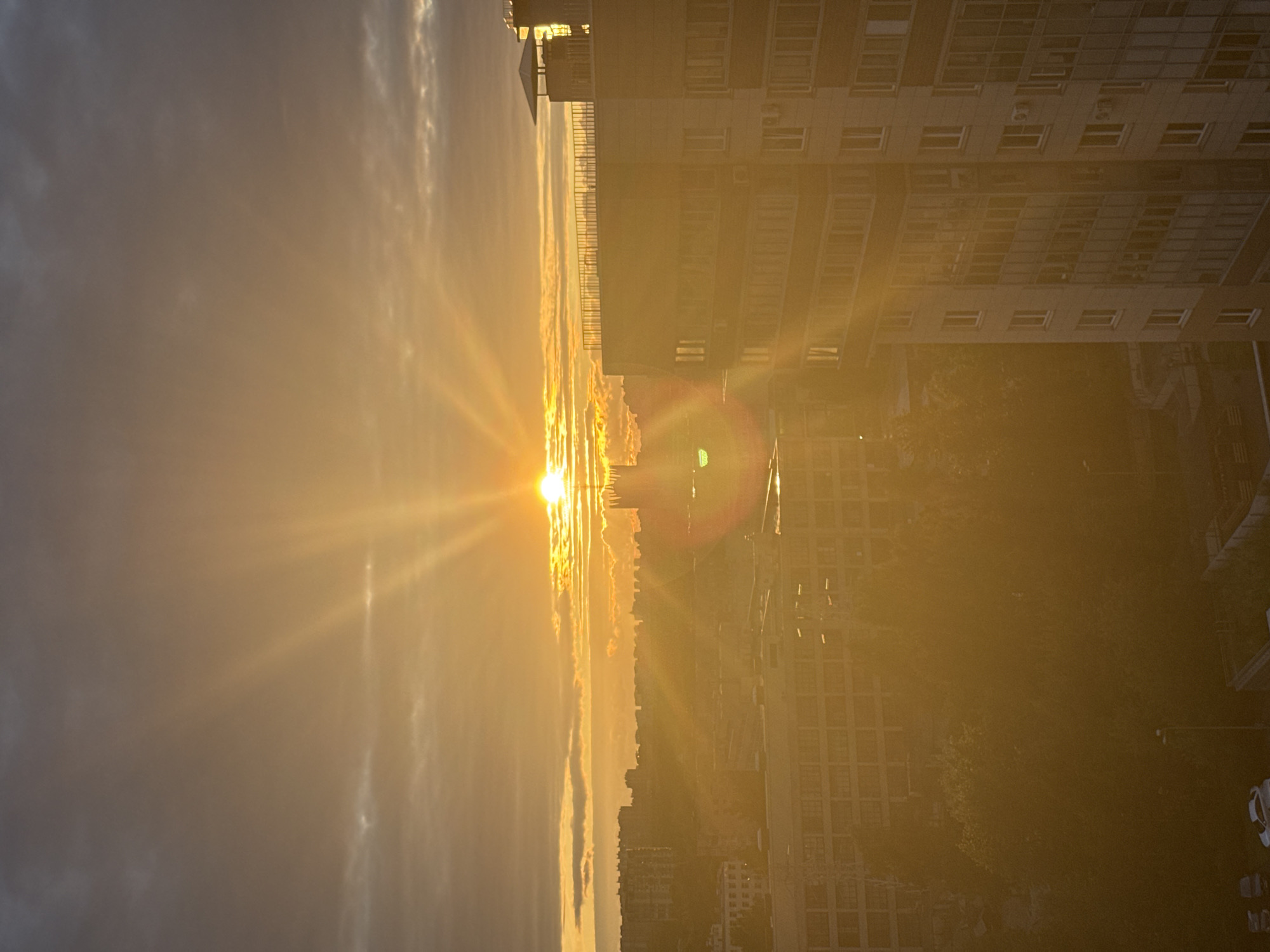 The image captures an urban landscape during a sunset. The sun is a bright orb in the upper center of the frame, casting golden rays that stretch across the sky and illuminate the scene. Clouds are visible, with some tinged orange by the setting sun. In the foreground, a modern apartment building dominates the right side of the image, its windows reflecting the warm light. To the left and in the background, other buildings of various sizes and architectural styles are visible, creating a dense urban environment. Trees are present in the middle ground, their foliage appearing dark in contrast to the bright sky. A few cars are parked in the lower left corner of the image. The overall impression is one of a city bathed in the warm glow of sunset. The scene is a typical urban view, likely from an apartment window, overlooking a residential area.