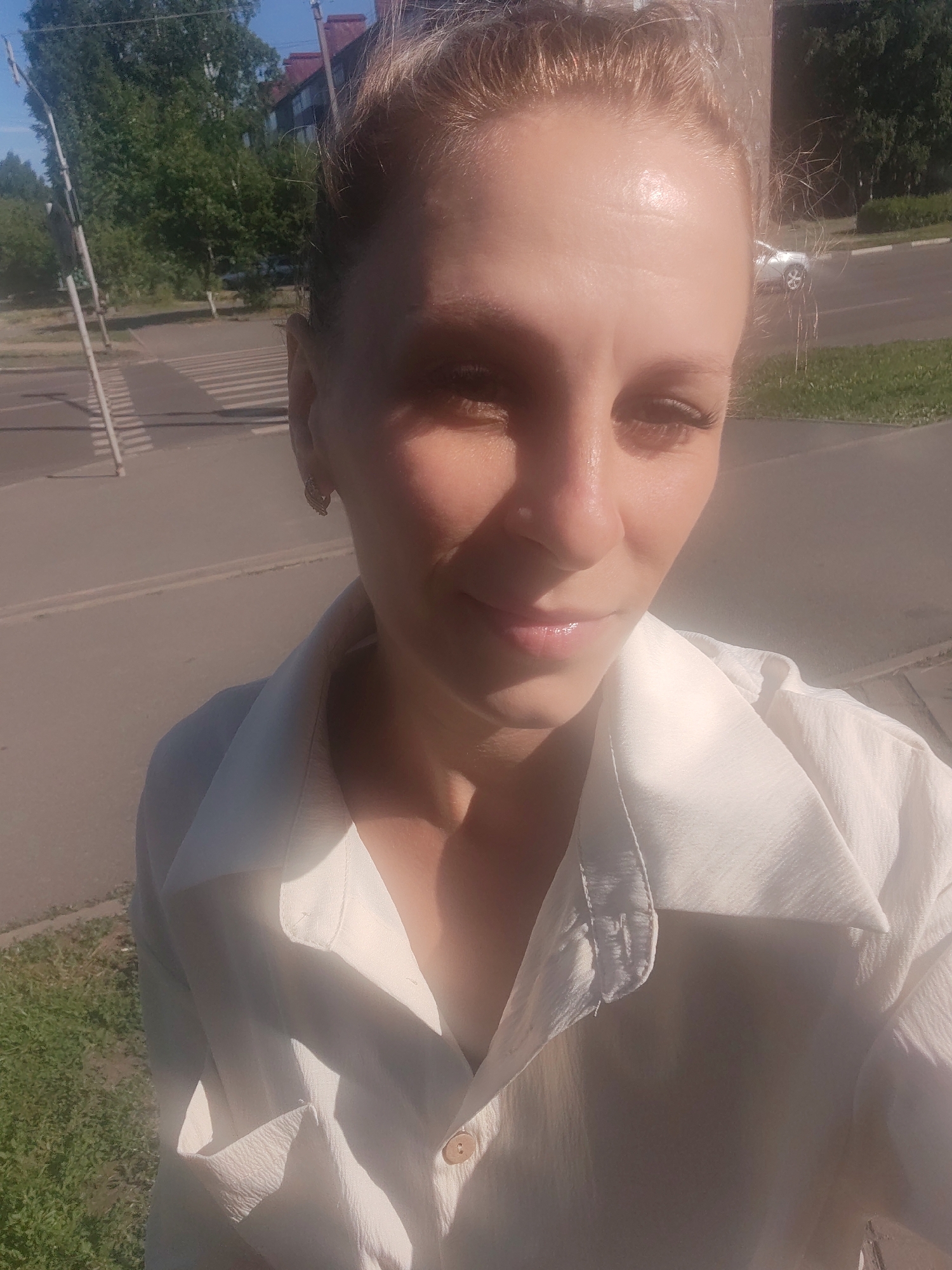 A woman with blonde hair, wearing a white shirt, poses for a selfie on a sunny day. The background shows a street with a crosswalk, trees, and a building. There are cars visible on the street. The image appears to be taken outdoors in an urban setting. The woman is looking directly at the camera. The lighting suggests it is daytime, likely afternoon given the angle of the sun.