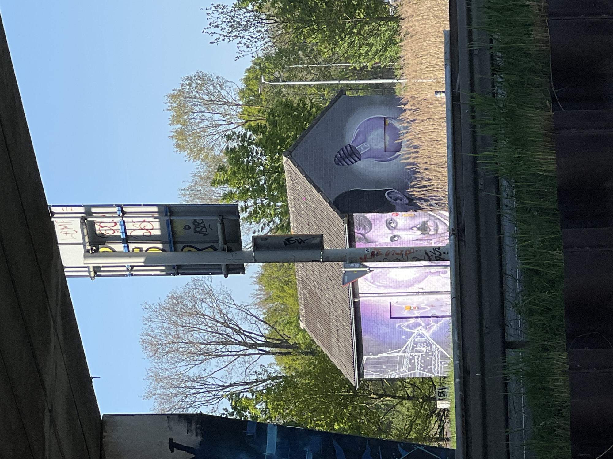 This media file captures an outdoor scene, likely beneath or adjacent to a bridge or overpass, featuring a small building adorned with vibrant graffiti murals. The image is taken on a clear, sunny day, indicated by the bright blue sky and distinct shadows.
The central subject is a single-story building with a dark-tiled roof. Its walls are covered in eye-catching street art. On the upper portion of the wall, a large, glowing purple light bulb is depicted, radiating a bright yellow-orange light from its filament. Below this, a portion of a human-like face, rendered in purple and grey tones, is visible, showing eyes and the bridge of a nose. Further down the wall, another mural features a white outline drawing of a house or building, surrounded by what appears to be lightning bolts or electrical arcs, also in purple hues.
Above the graffitied building, a large, grey metallic sign structure is prominent, partially covered with various illegible tags and some discernible red letters that seem to spell "AMF" or similar. To the left, a concrete structure, possibly the underside of a bridge, frames the scene. In the background and foreground, numerous trees with green foliage are scattered, suggesting spring or early summer, though one tree to the left shows bare branches. On the far right, a vertical barrier or wall is partially covered with both dry, light-colored reeds and fresh green grass. No specific activity or event is taking place; the image is a static capture of urban art in an outdoor setting in Weert, Netherlands.