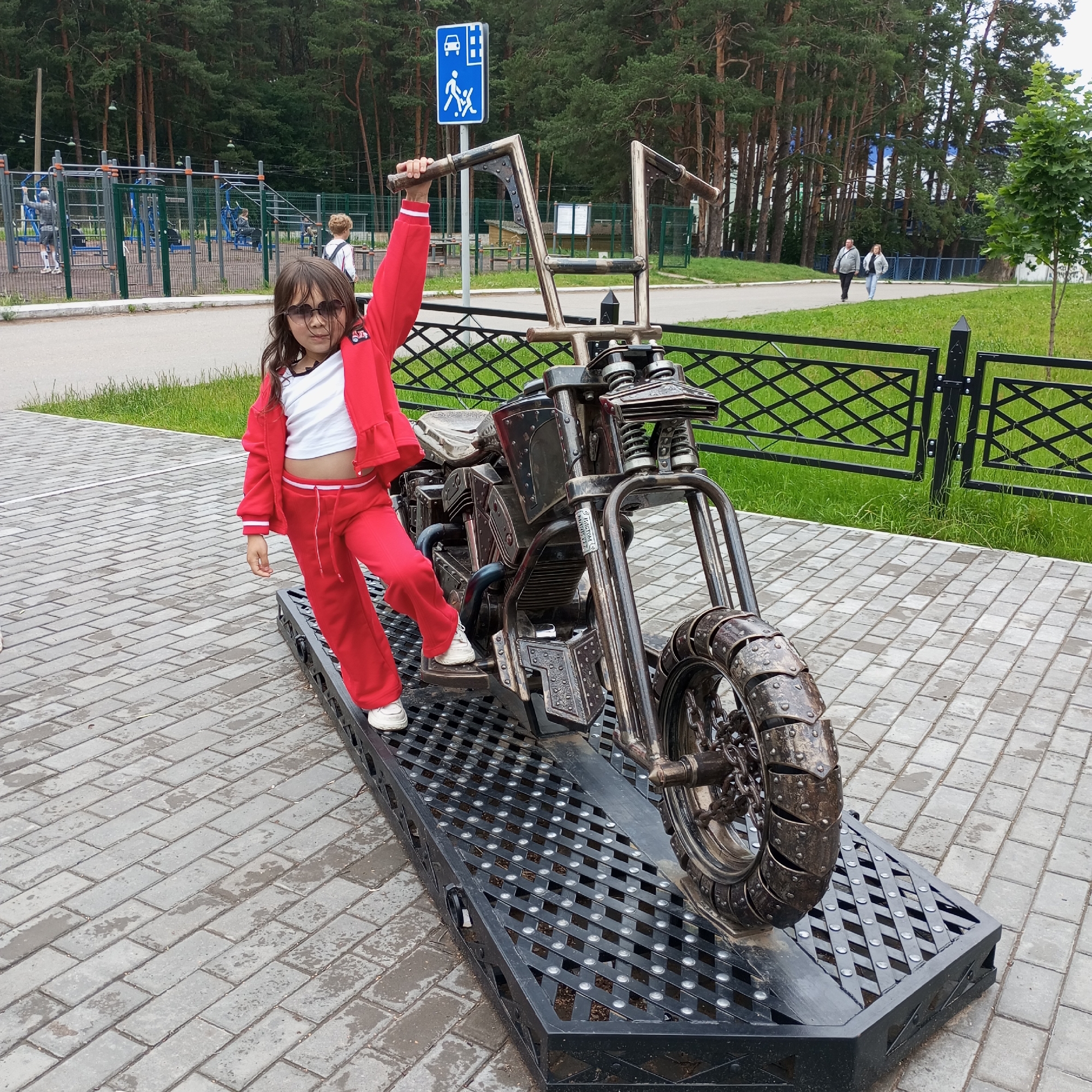 A young girl, wearing a red tracksuit and sunglasses, stands with her right arm raised, holding onto the handlebars of a metallic sculpture of a motorcycle. Her left foot is on the motorcycle's platform, and her right foot is on the ground. The motorcycle sculpture is elaborate and appears to be made of scrap metal, with intricate details.
The scene is set outdoors on a paved area, likely a park or public space, with a black metal fence and green grass in the background. Behind the fence, a row of tall pine trees forms a dense forest. In the distance, there are elements of a playground with blue and silver equipment and other people. A blue sign with white pictograms, depicting a car, a pedestrian, and a building, is visible to the left of the girl. Two adults are walking on a paved path in the background.
The ground is wet, suggesting it may have recently rained or is damp. The lighting appears to be overcast, indicating a cloudy day. The girl's posture and raised arm suggest a playful or triumphant pose, possibly posing for a photograph. No visible text other than on the blue sign is discernible in the image. The overall impression is a casual outdoor scene, likely in a public recreational area in Chaikovsky, Russia.