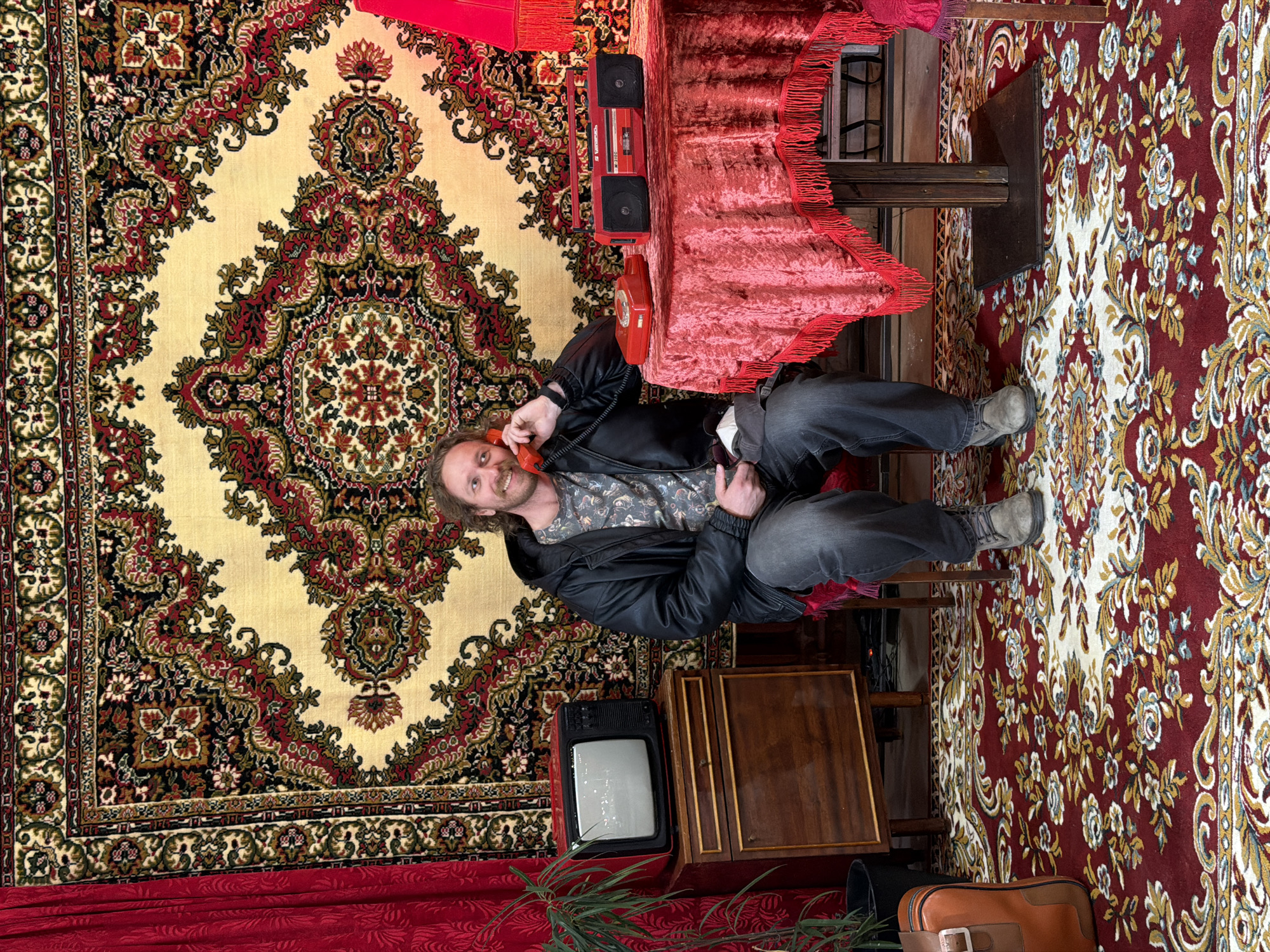 A Caucasian man with long brown hair sits in a cluttered room. He is wearing a black leather jacket, a patterned shirt, and dark jeans. He is holding a red rotary phone to his ear, smiling. The room is decorated with multiple patterned rugs, one on the wall behind him and another on the floor. To the left of the man is a small vintage television set, and next to it, a wooden cabinet. A table covered with a red fringed tablecloth and a red boombox is behind him. A brown suitcase is in the foreground. The image evokes a sense of retro or kitsch decor, possibly simulating a Russian interior from the Soviet era. It is likely a scene from a play, movie, or themed restaurant/bar. Given the details of decor, the photo is possibly taken in Saint Petersburg, Russia.