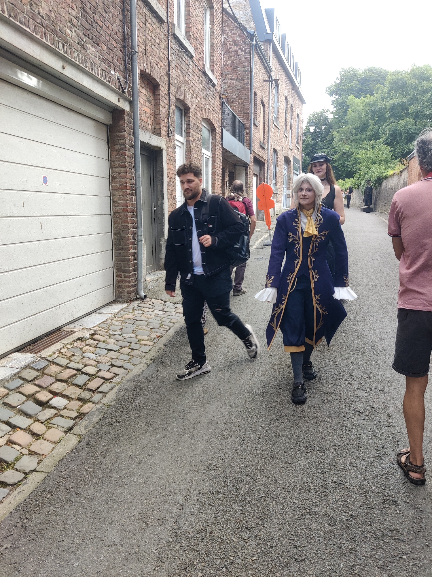 In a narrow alleyway, likely in the city of Marche-en-Famenne, Belgium, several individuals are walking on a cobblestone and asphalt surface. The alley is flanked by brick buildings, one with a white garage door. In the foreground, a man with curly hair, wearing a black jacket over a white t-shirt and black jeans, walks to the right. Behind him, a person in a striking blue and gold costume with a white ruffled collar and cuffs is walking with a neutral expression, accompanied by another person in black. Further down the alley, other figures are visible, including one with a red backpack and another person in a pink shirt and dark shorts on the far right. The overall scene suggests an outdoor event or gathering taking place in an urban environment.