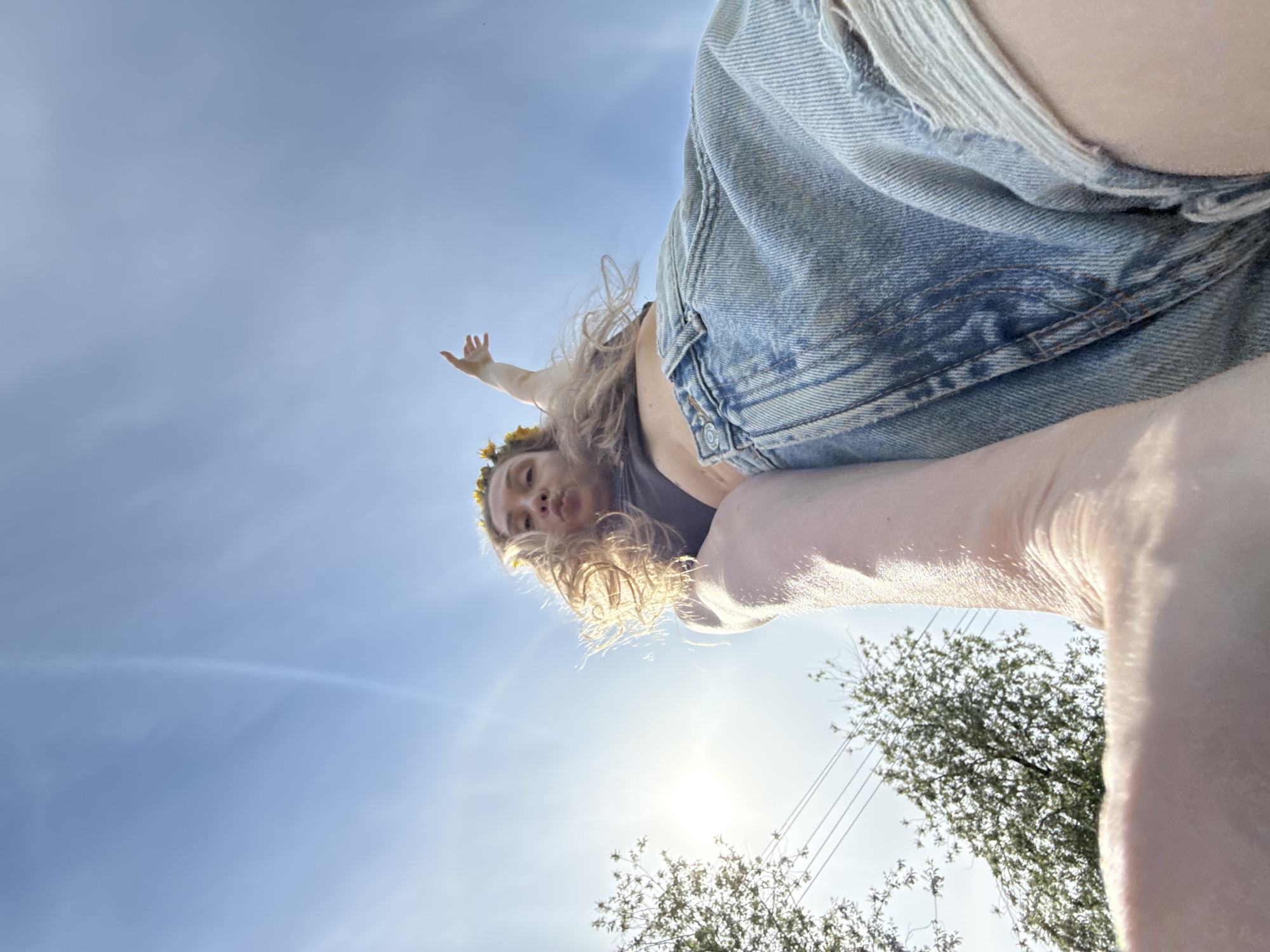 A low-angle shot depicts a person wearing denim shorts and a floral crown, positioned against a blue sky. The sun is directly behind the person, causing lens flare. A hand is raised in the upper left. Trees are visible at the bottom right. There is no indication of the location being Nizhnyaya Chermoda, Russia.