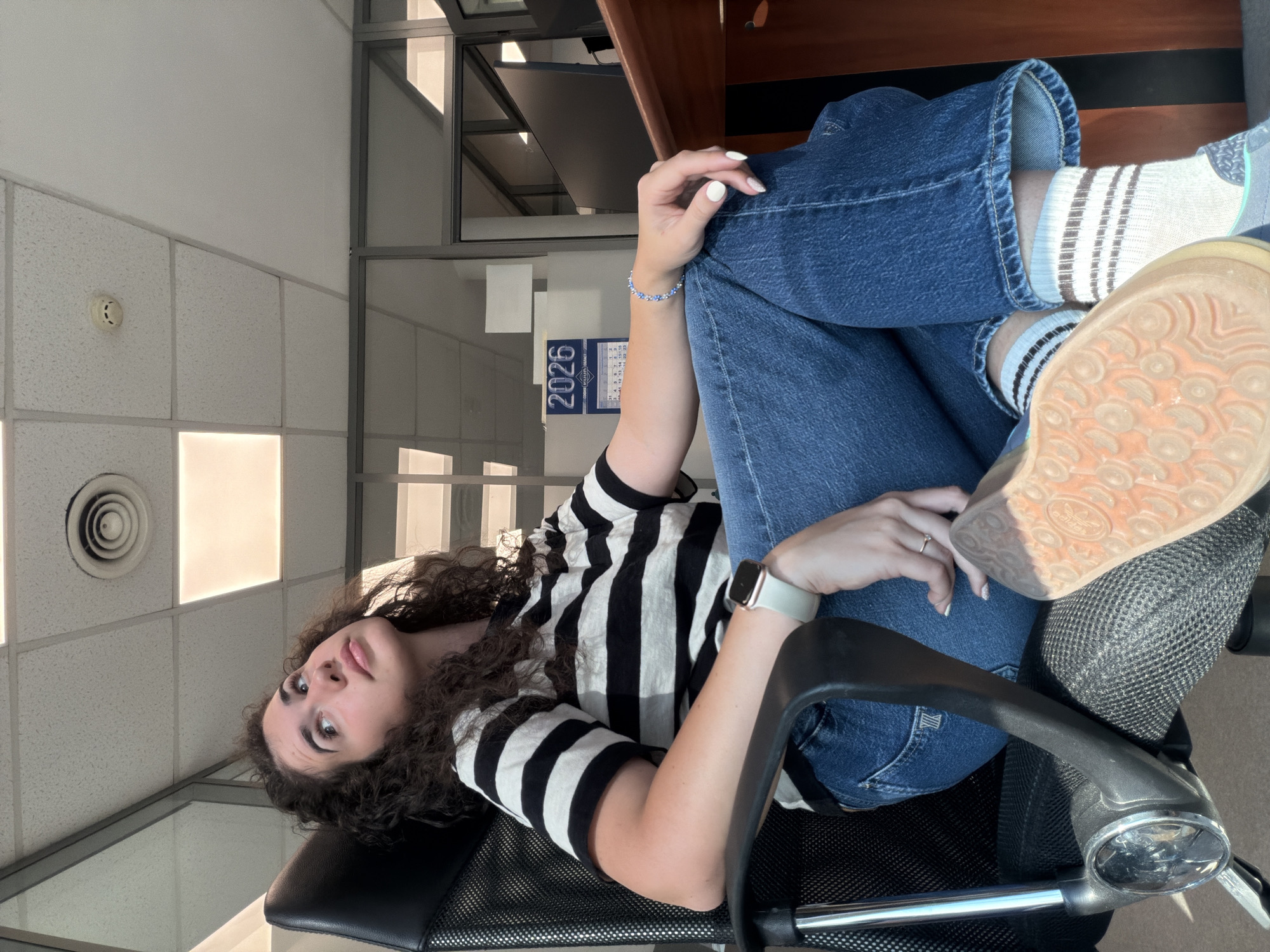 A young woman with curly dark hair is seated in a black office chair, facing left. She is wearing a black and white striped t-shirt, blue denim jeans, and white socks with brown stripes, along with grey and blue sneakers. Her left leg is crossed over her right, with the sole of her left shoe visible. She is looking upwards and to the left, with her right hand resting on her left knee. She is wearing a white smartwatch on her left wrist and a beaded bracelet on her right wrist. A ring is visible on her right ring finger. In the background, there is a desk with a computer monitor, a calendar displaying "2026," and glass partition walls. The ceiling has fluorescent lighting and a circular air vent. The image appears to be taken indoors, possibly in an office environment in Sofia, Bulgaria.