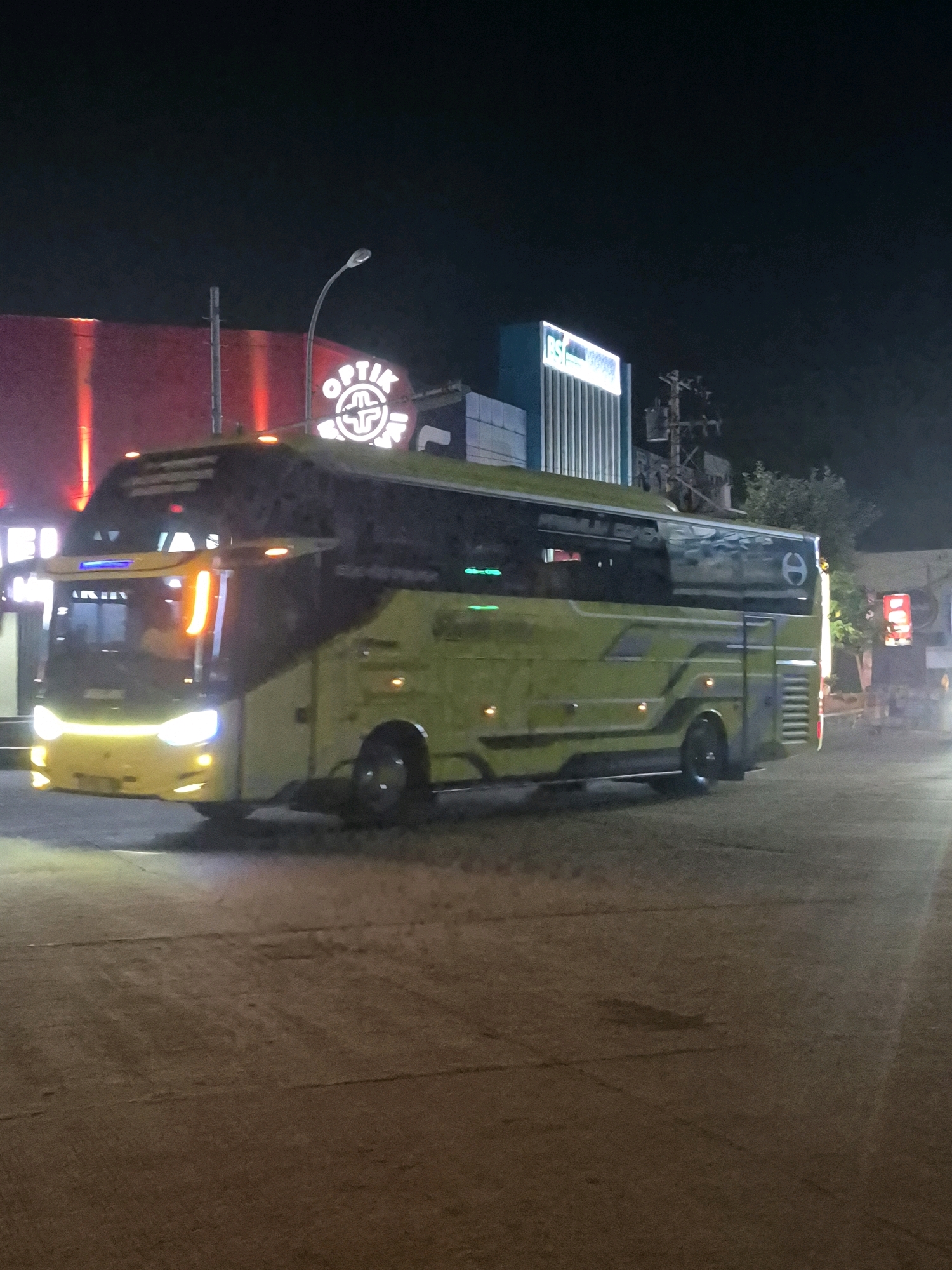 This image captures a yellow bus at night on a street in Surakarta, Indonesia. The bus, a large passenger vehicle, is parked or slowly moving on a paved road. Its headlights and other lights are illuminated. In the background, several buildings are visible with illuminated signs. One sign prominently displays "OPTIC" in large, glowing letters, suggesting a retail establishment. Another sign, to the right of the "OPTIC" sign, has a blue and white color scheme and appears to be part of a different building. The overall lighting suggests it is nighttime, with artificial lights from the buildings and the bus creating illumination. The environment appears to be an urban setting, likely a commercial area given the presence of retail signage. The road surface is visible in the foreground and shows some texture and markings.